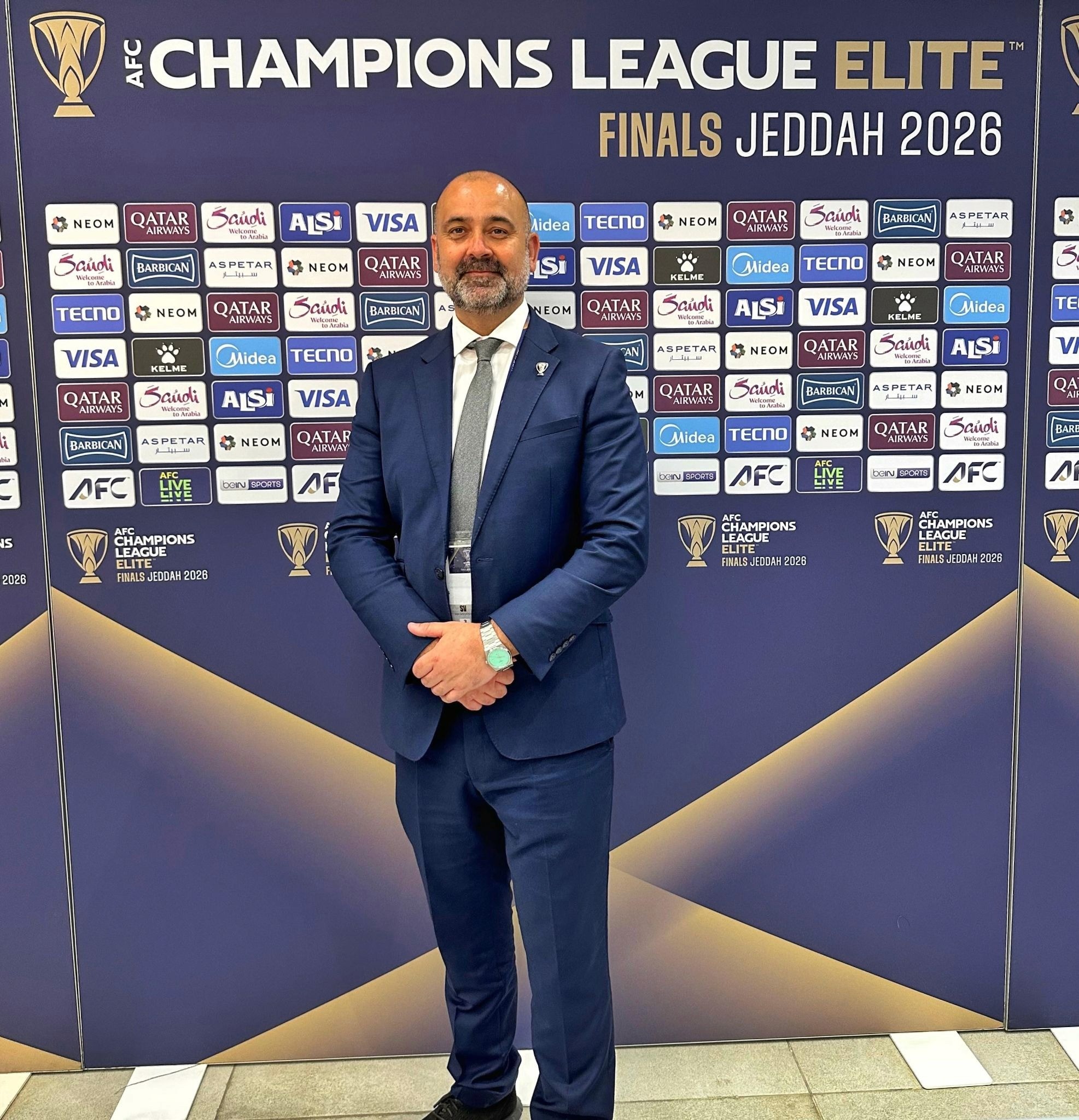Man in blue suit posing at AFC Champions League Elite Finals Jeddah 2026 media wall.
