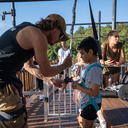 在一个木制平台上，一位教练正在帮助一名儿童调整攀岩装备，背景中两位女士和其他人正在等待。