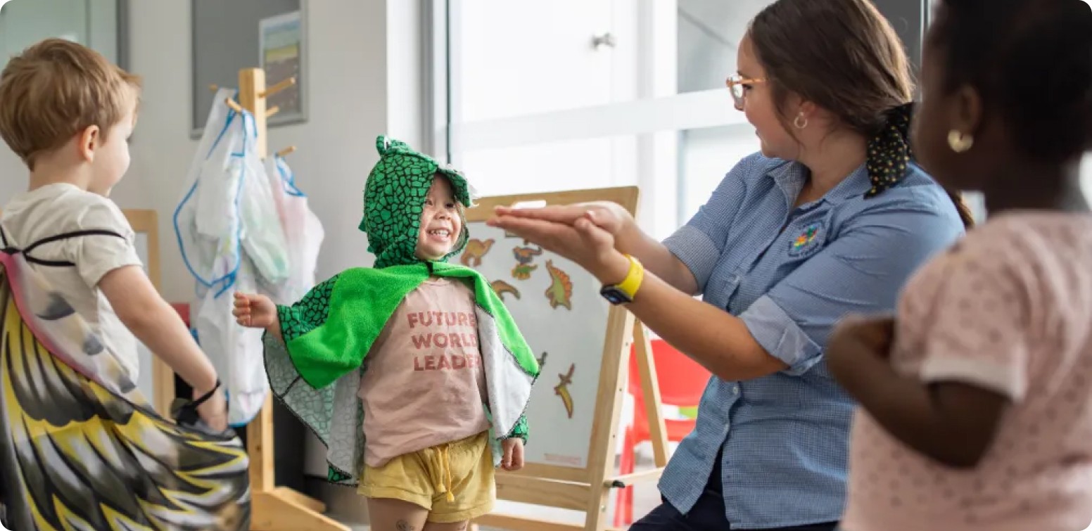 Educator helping a smiling child dress up in a green dinosaur costume at Kaleidoscope Kids Early Learning Centre, encouraging play-based learning in a bright classroom.