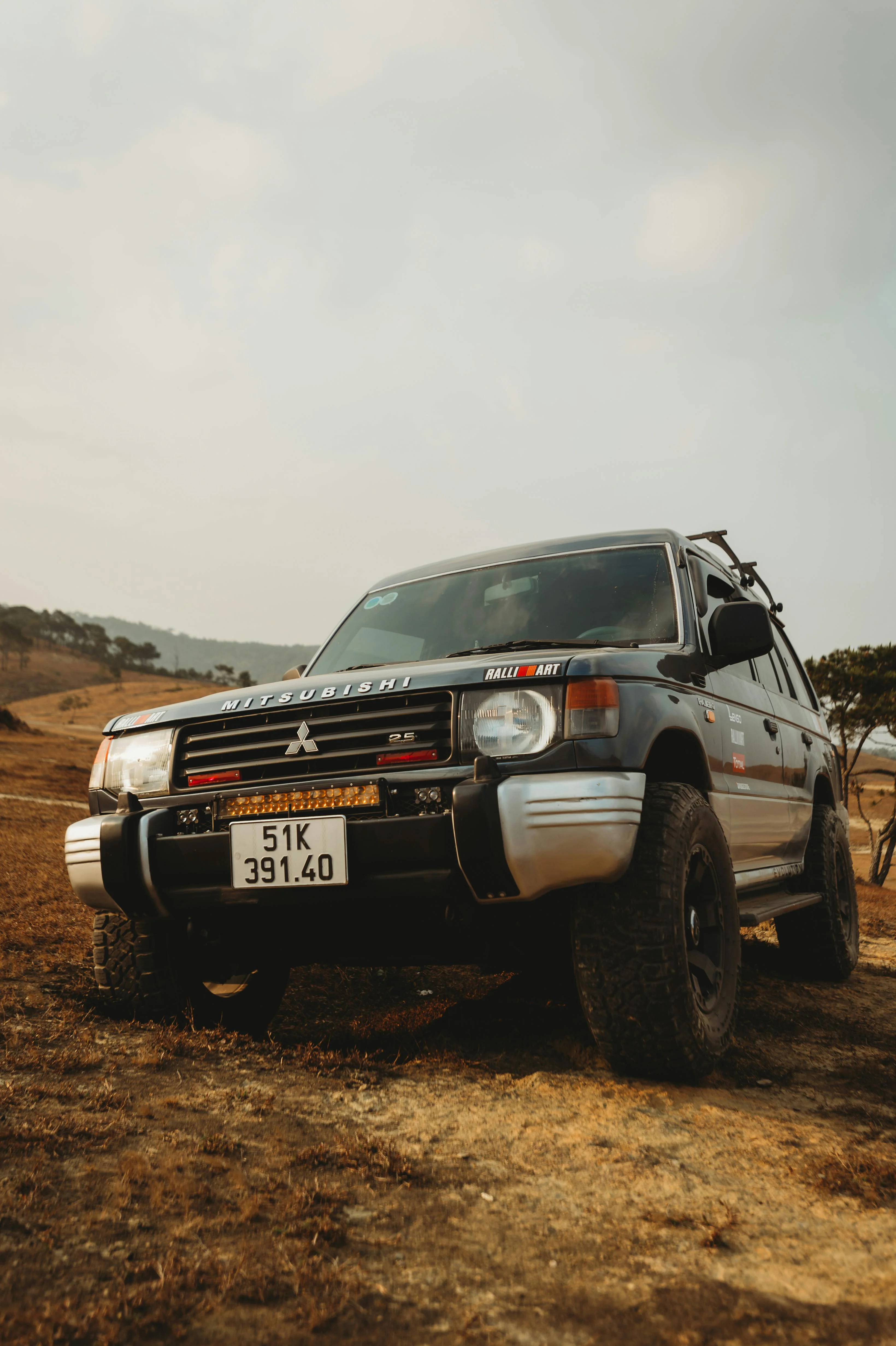 Gros plan sur un Mitsubishi Pajero équipé d’accessoires Ralliart et photographié sur un sol sec et rocailleux. Le 4x4 japonais arbore une préparation baroudeuse avec pneus tout-terrain, protections renforcées et éclairage additionnel, rappelant son héritage mythique au Dakar. L’arrière-plan de collines et le ciel pâle accentuent l’impression d’aventure brute et d’endurance mécanique, soulignant le caractère robuste et indestructible du Pajero.
