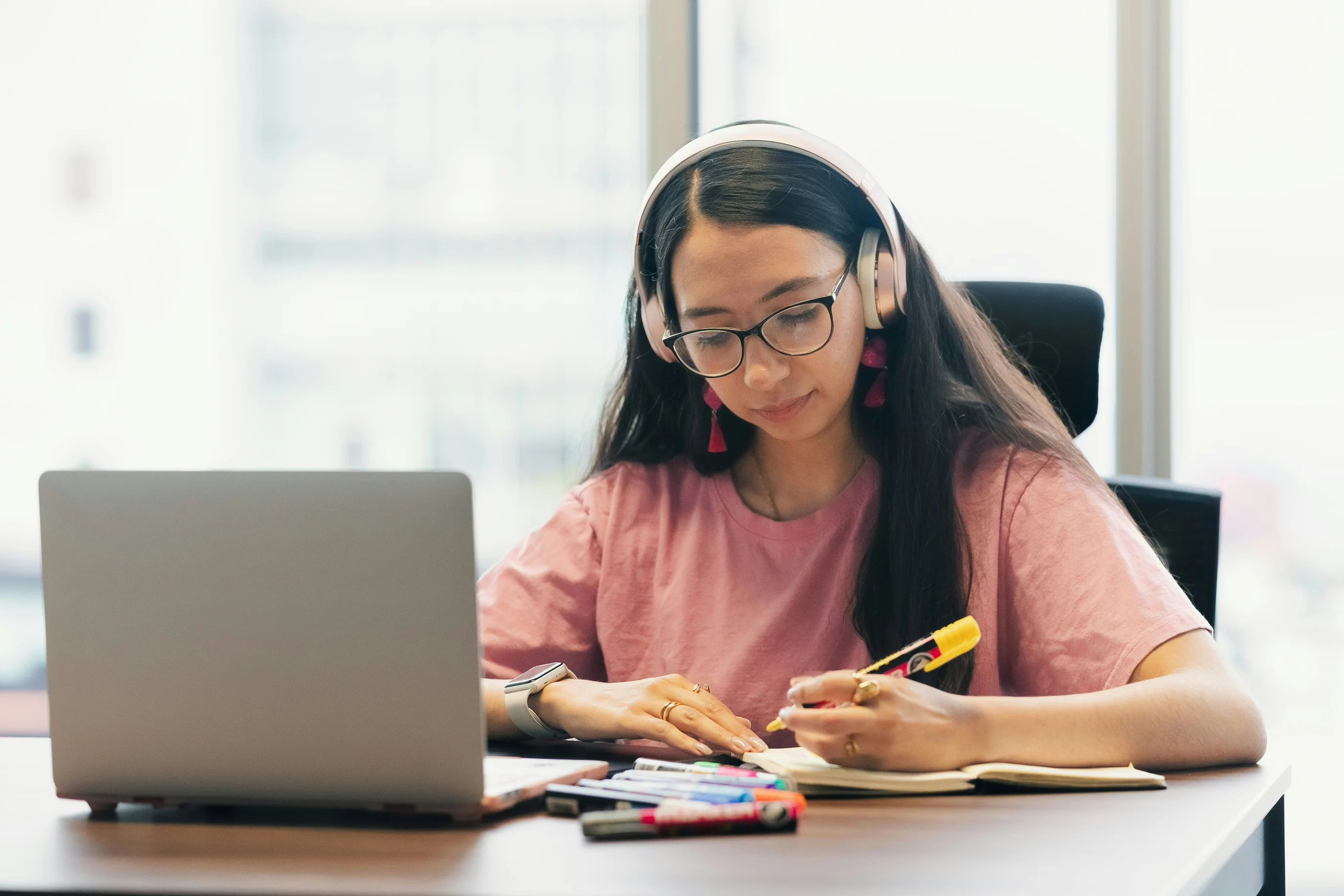 A woman wearing headphones and taking notes beside her laptop.