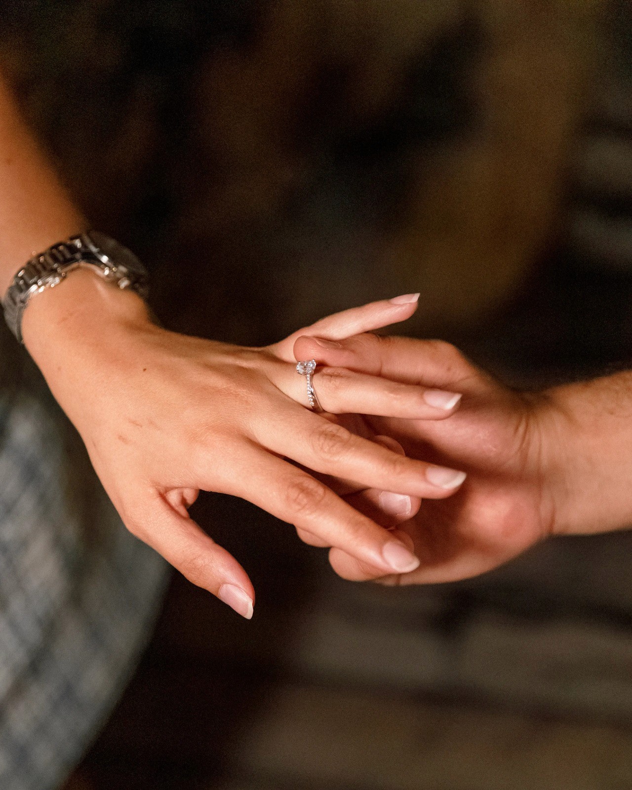 Close-up of two hands gently clasped, one wearing a sparkling engagement ring. The soft lighting and pastel nails evoke a sense of romance and intimacy.