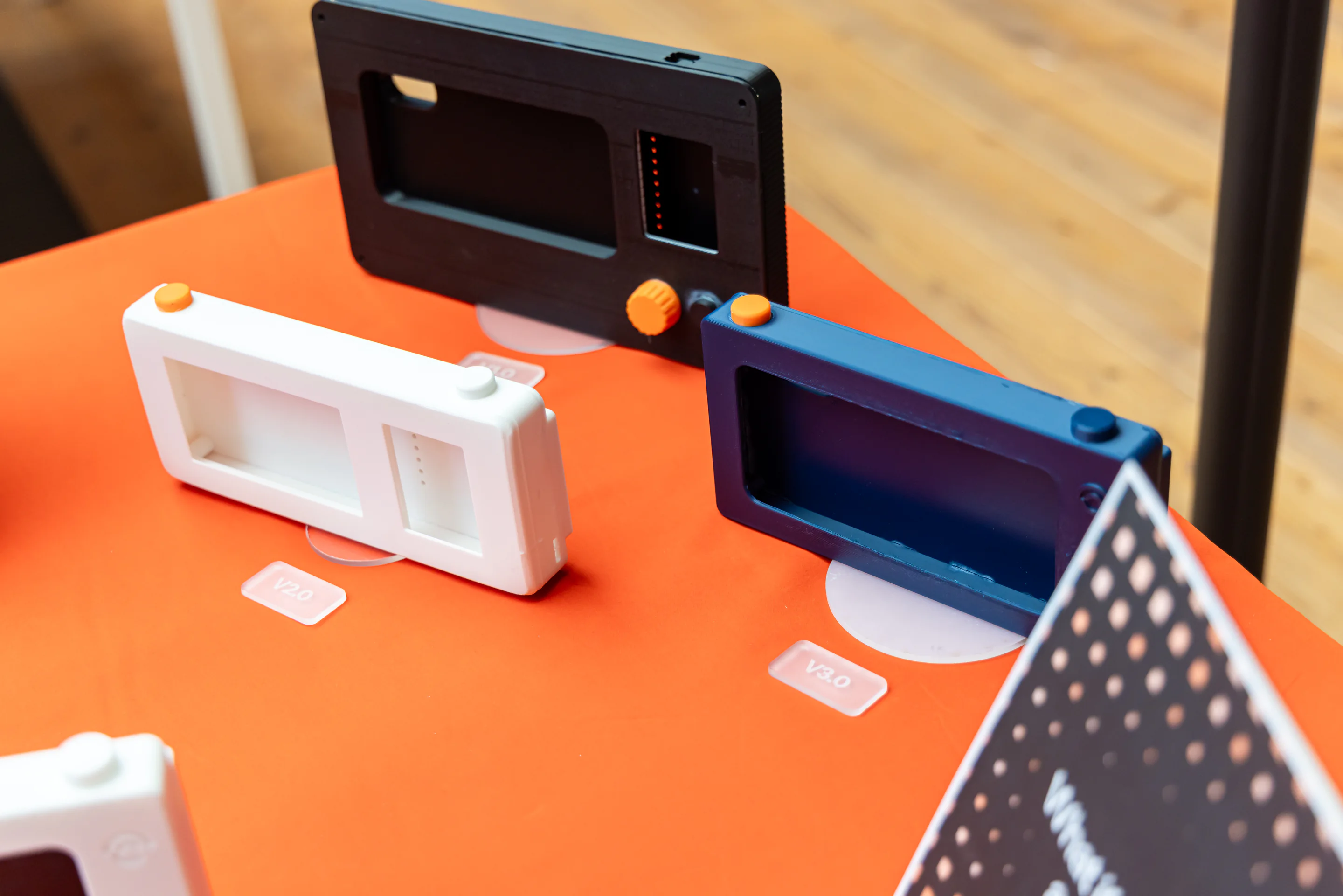 Three rectangular camera enclosure prototypes—white, black, and dark blue—stand on an orange exhibition table.