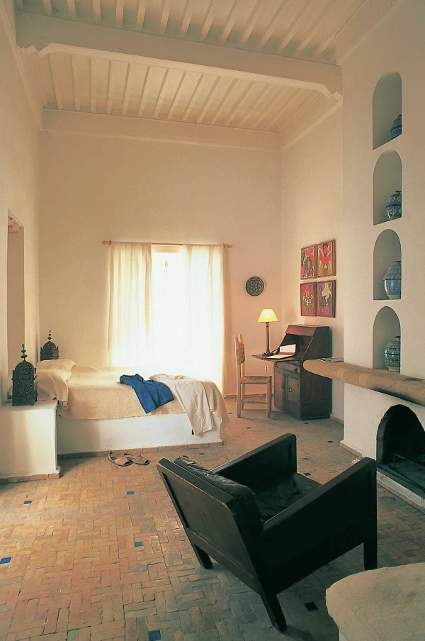 Warm morning light fills a whitewashed room with light linen curtains, a vintage wooden writing desk and banked stone fireplace.