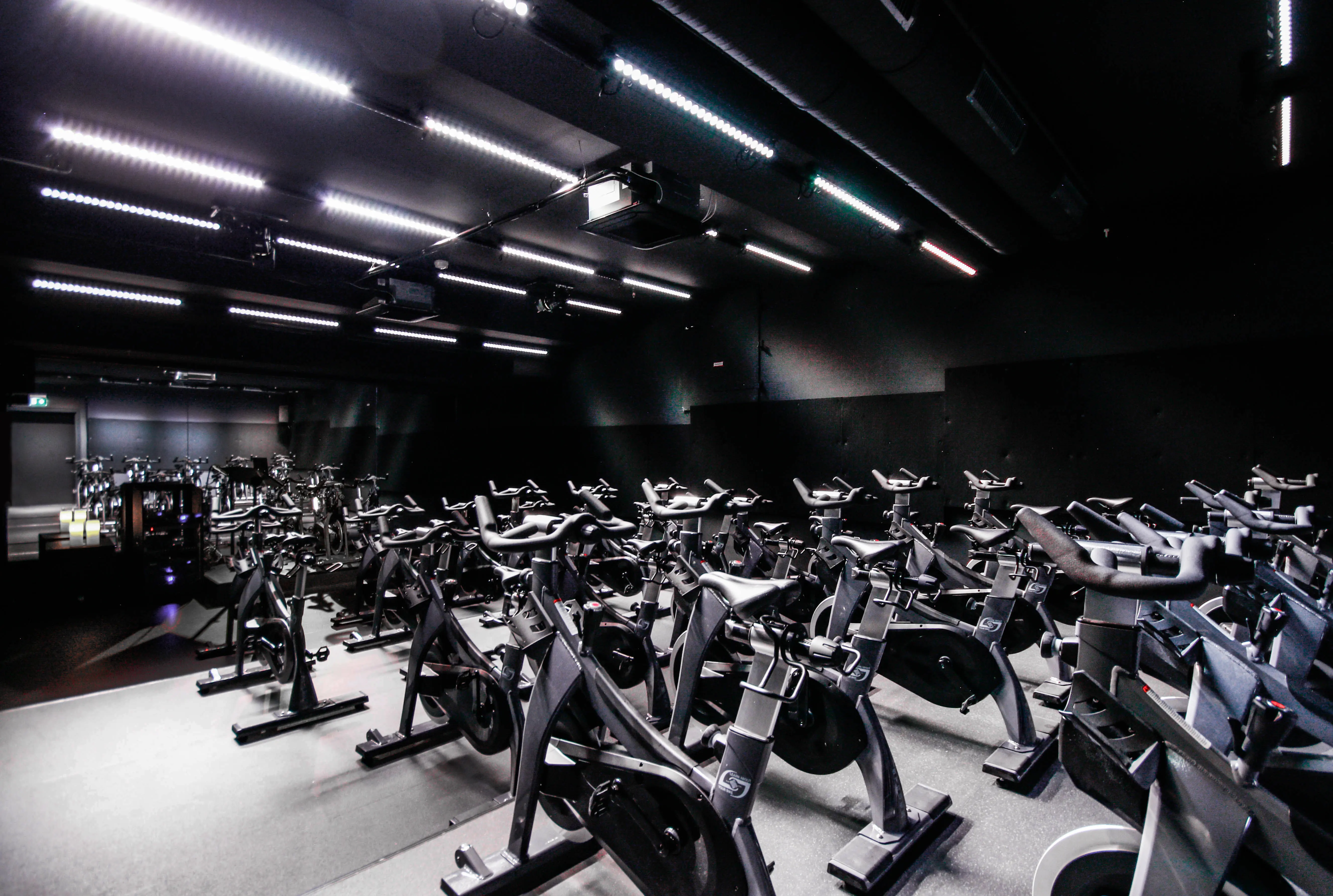 Dark spinning studio at CYCLA with rows of black spin bikes under linear white lights on a black ceiling.