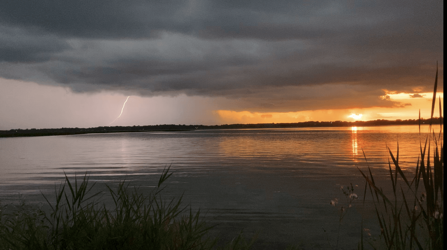Lightning flashing over a lake as storm clouds fade into a golden sunset.