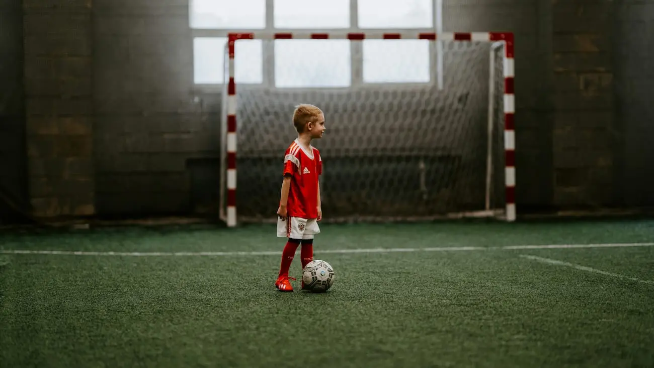 A young junior player in a red jersey stands on an indoor football pitch in front of a mini goal, ready to play as part of a mini football league that emphasises and supports the fun and fundamentals of football for children.