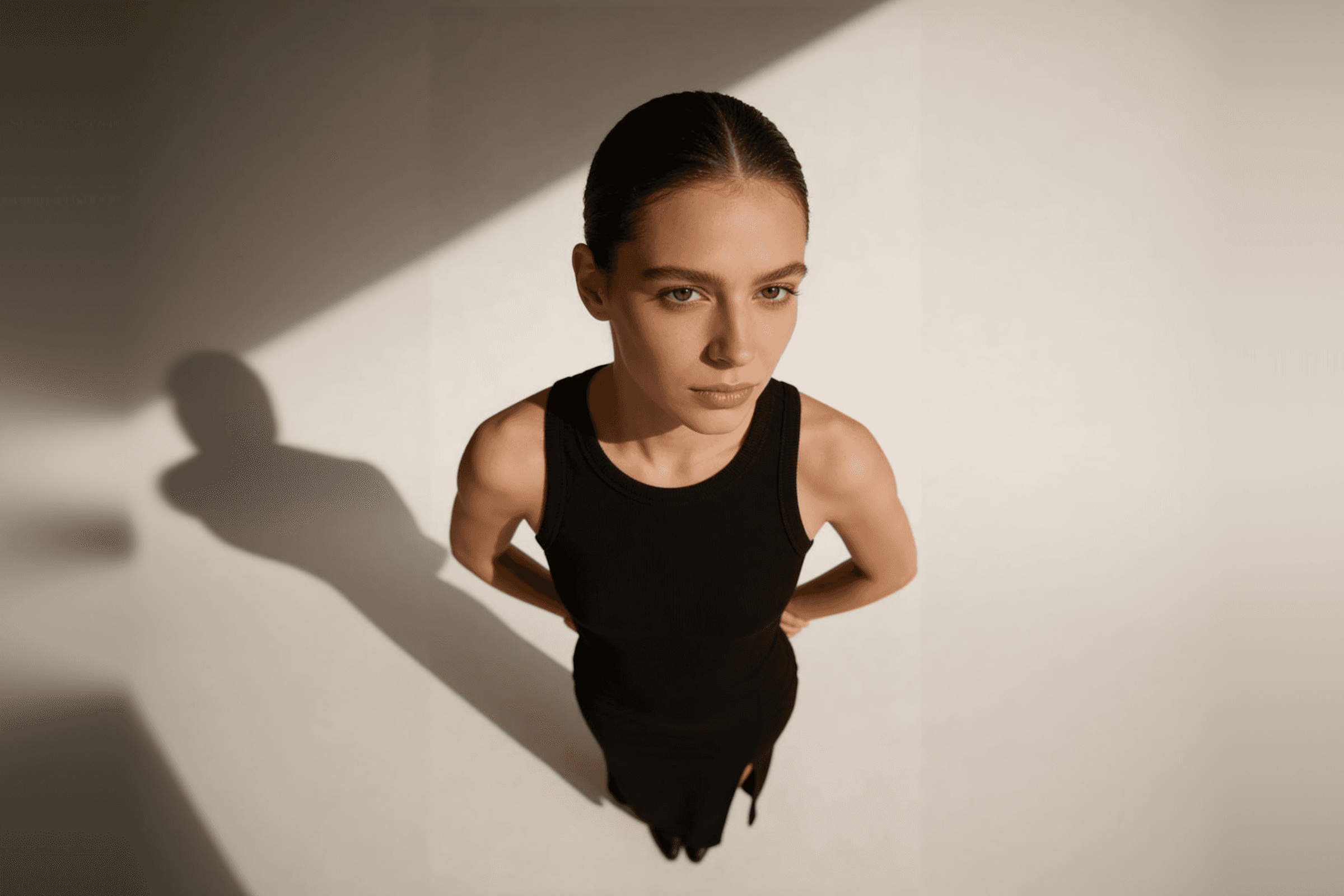 A woman in a black sleeveless dress stands confidently in soft lighting, her hair tied back and her expression calm and focused.