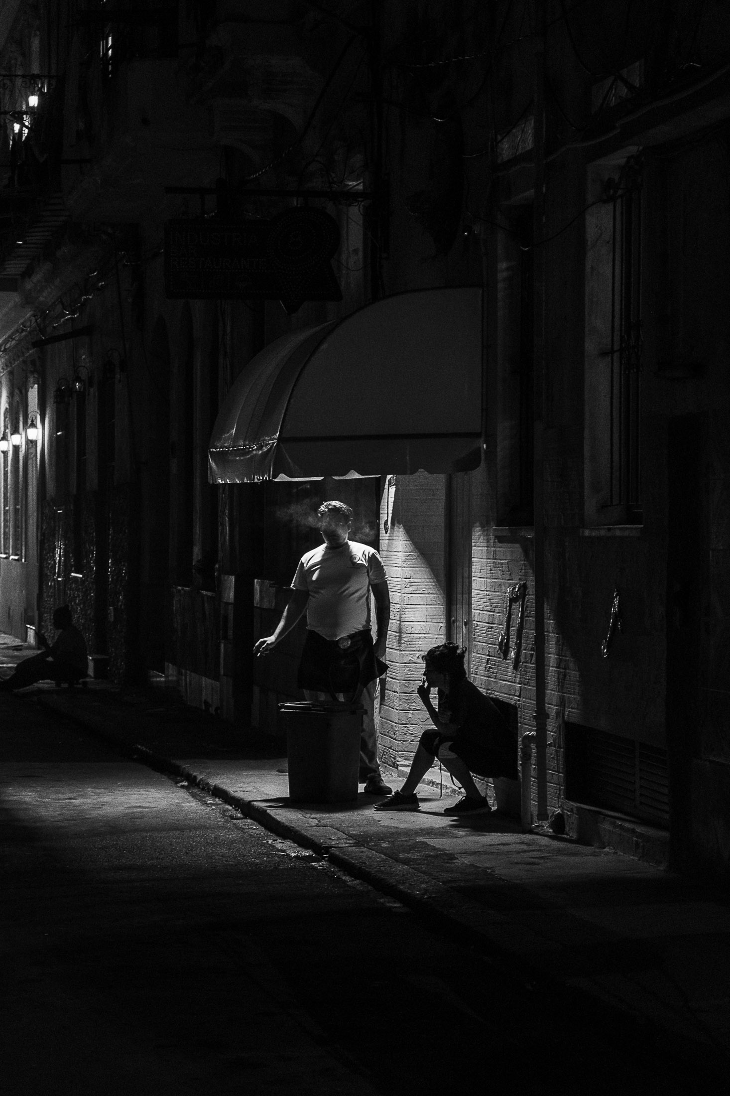 Black and white photograph of a man smoking under a street light while a child sits nearby in a dark alley, street photography by Richard Peterson.