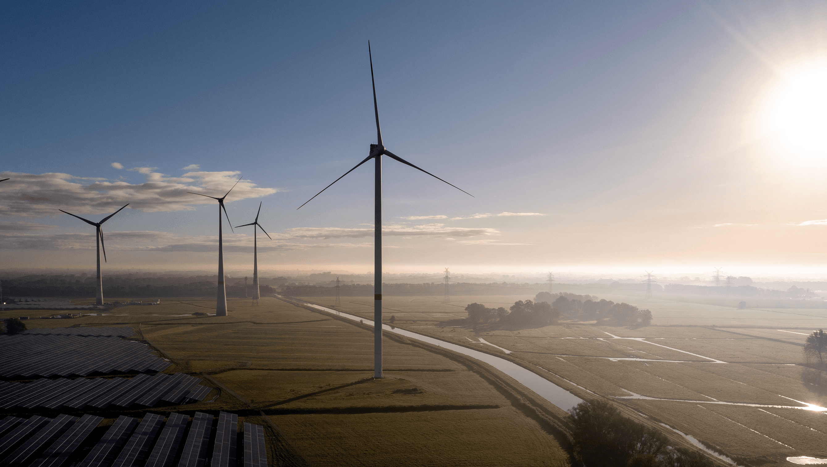 Windkraftanlagen stehen in einer weiten Landschaft und werden von sanftem Sonnenlicht vor einem klaren blauen Himmel erleuchtet.