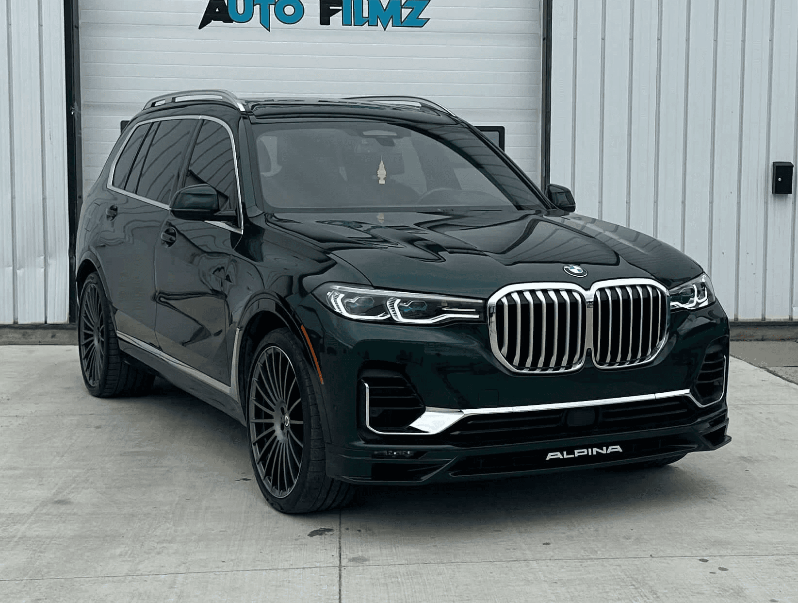 A black BMW Alpina XB7 luxury SUV is parked on a concrete driveway in front of a white industrial building.