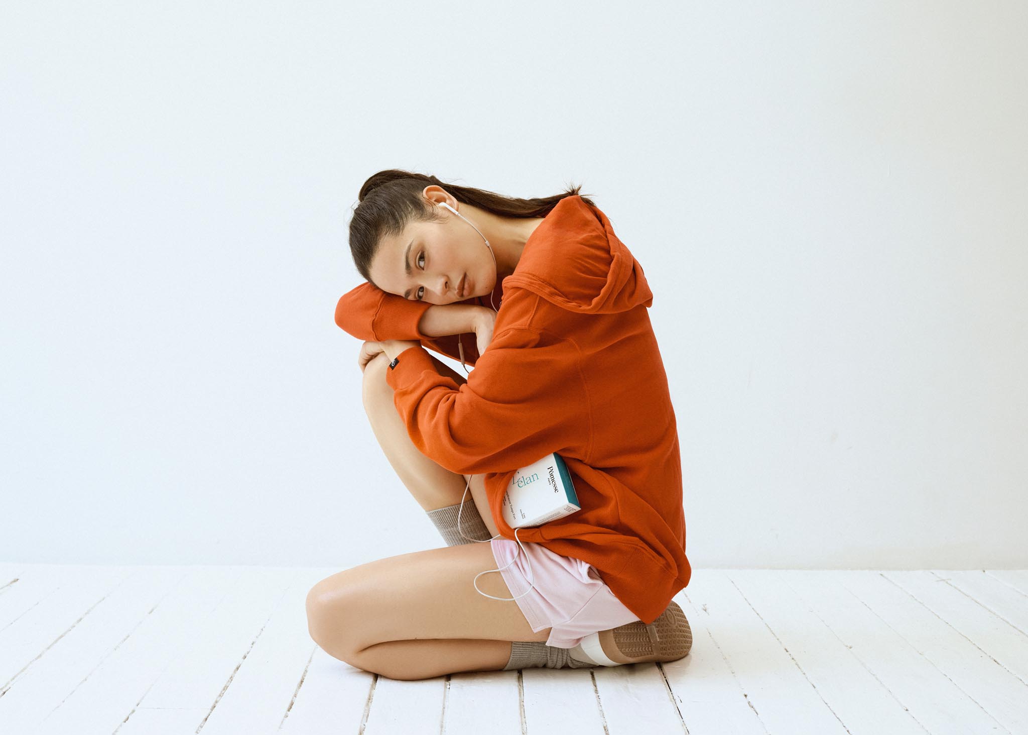 Femme assise au sol, portant un sweat orange et un short clair, tenant un produit Pômesse Paris contre elle
