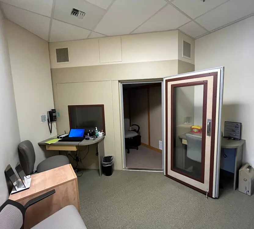 A small office space featuring a desk with a computer, an open door, and a soundproof booth in the corner.