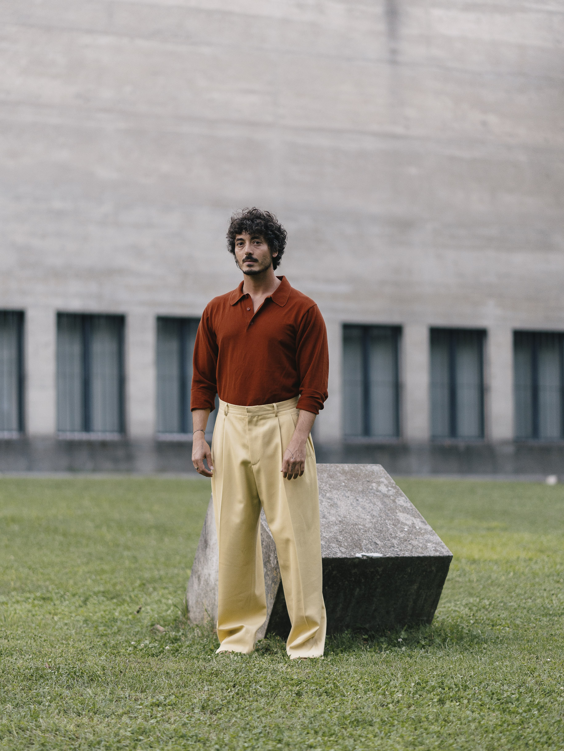 Portrait de l'artiste italien Lumi portant une chemise rouge retroussée et d'un pantalon jaune devant la bibliothèque cantonale de Lugano