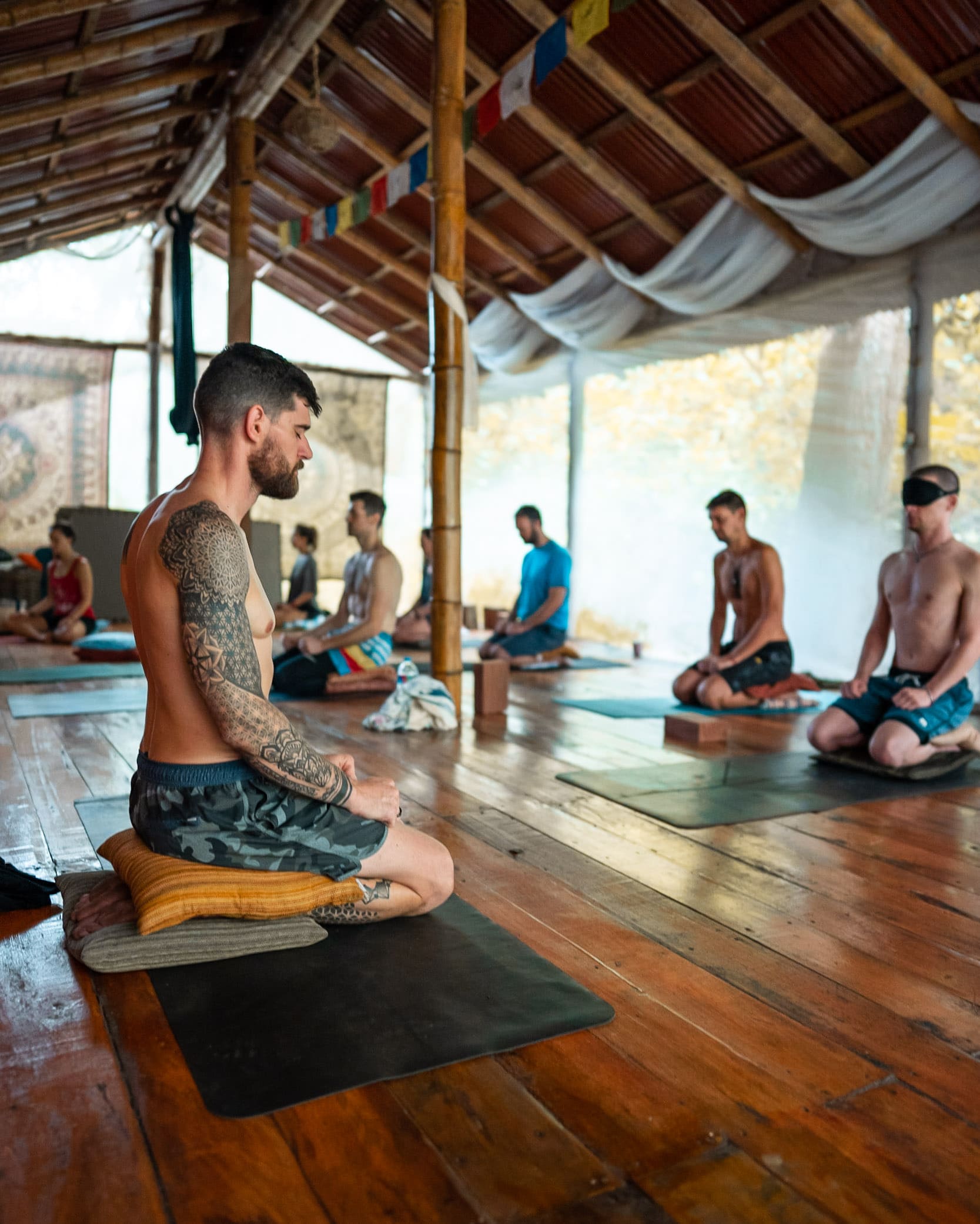 Group meditation session led by Flo in a serene room with wooden floors and bamboo pillars. People sit on yoga mats, with eyes closed, creating a calm atmosphere.