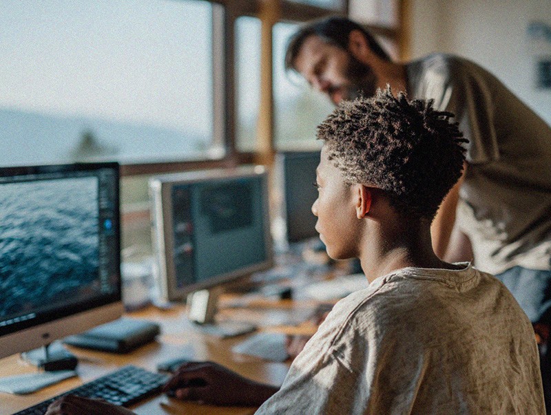 A young person with short, textured hair sits at a desk working on a desktop computer, focused on the screen. An adult stands slightly behind and to the side, leaning in as if offering guidance or instruction. The workspace includes multiple monitors, a keyboard, and other desk items, with large windows in the background letting in natural light, suggesting a classroom or learning environment.