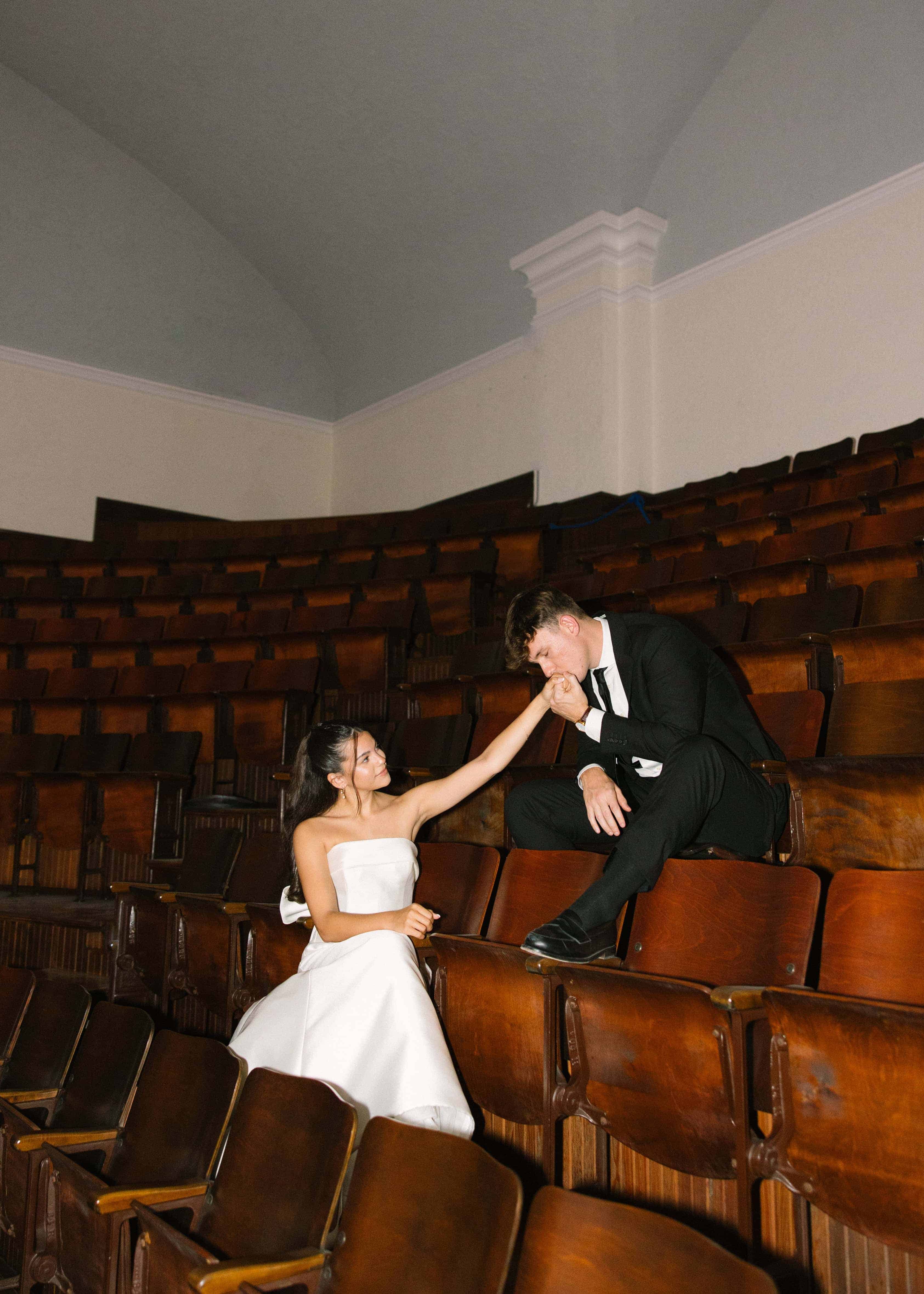 A man in a tuxedo is kneeling and kissing the hand of a woman in a white dress. They are in a room with rows of wooden chairs.