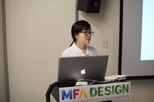 A photo of a woman giving a lecture from a laptop put on a stand with text MFA Design on it.