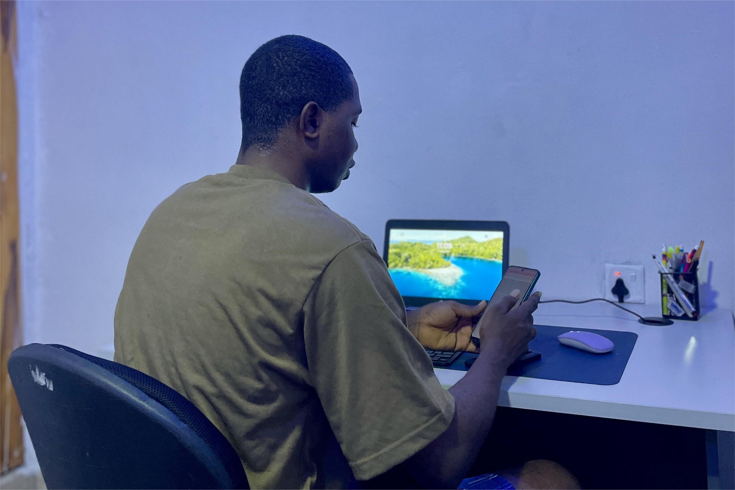 A person wearing headphones sits at a desk with a large curved monitor displaying graphic design software, surrounded by a laptop, smartphone, and potted plant, creating a modern workspace setup.
