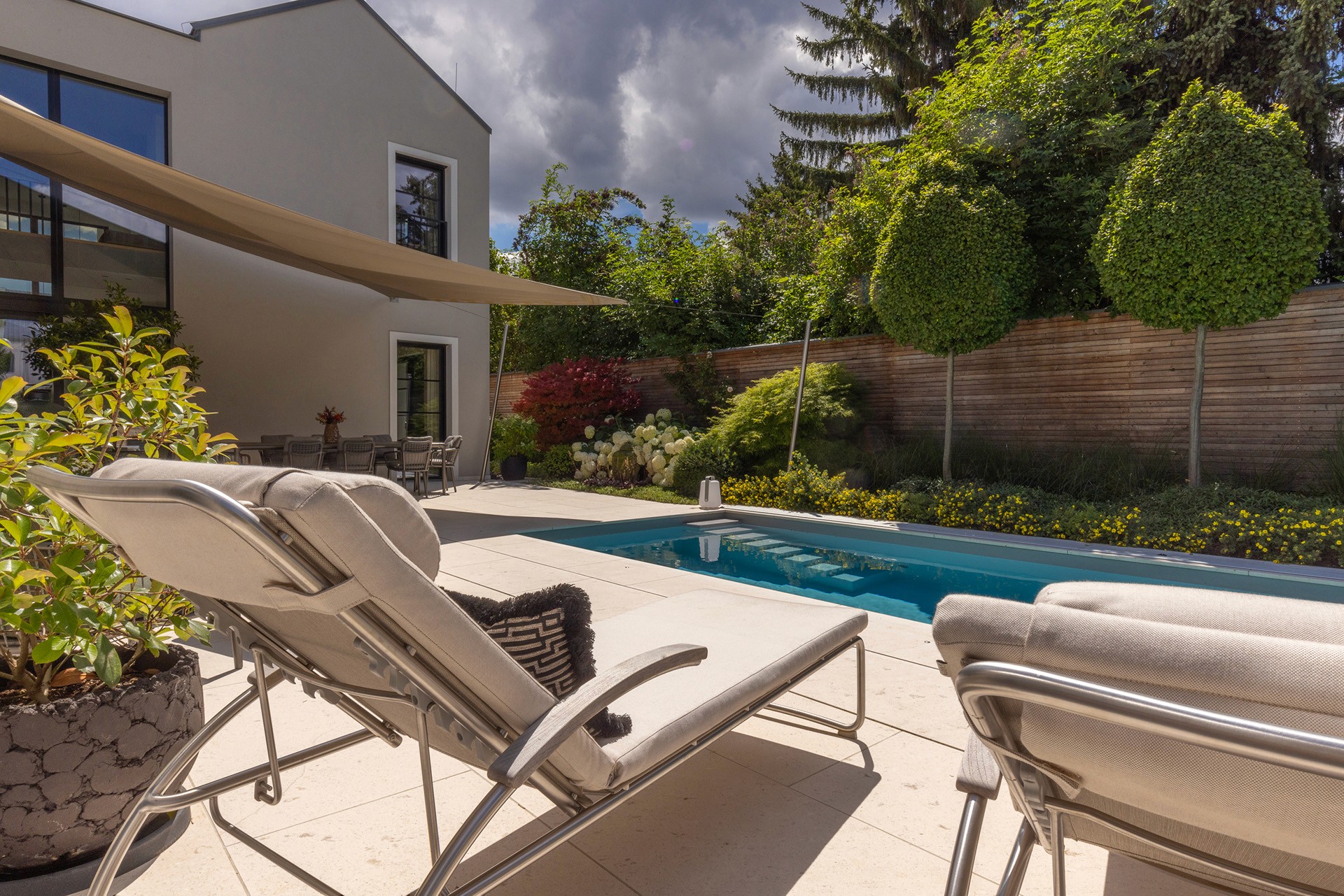 Moderne Poolterrasse mit Liegestühlen, Sonnensegel und strukturierter Gartenbepflanzung.
