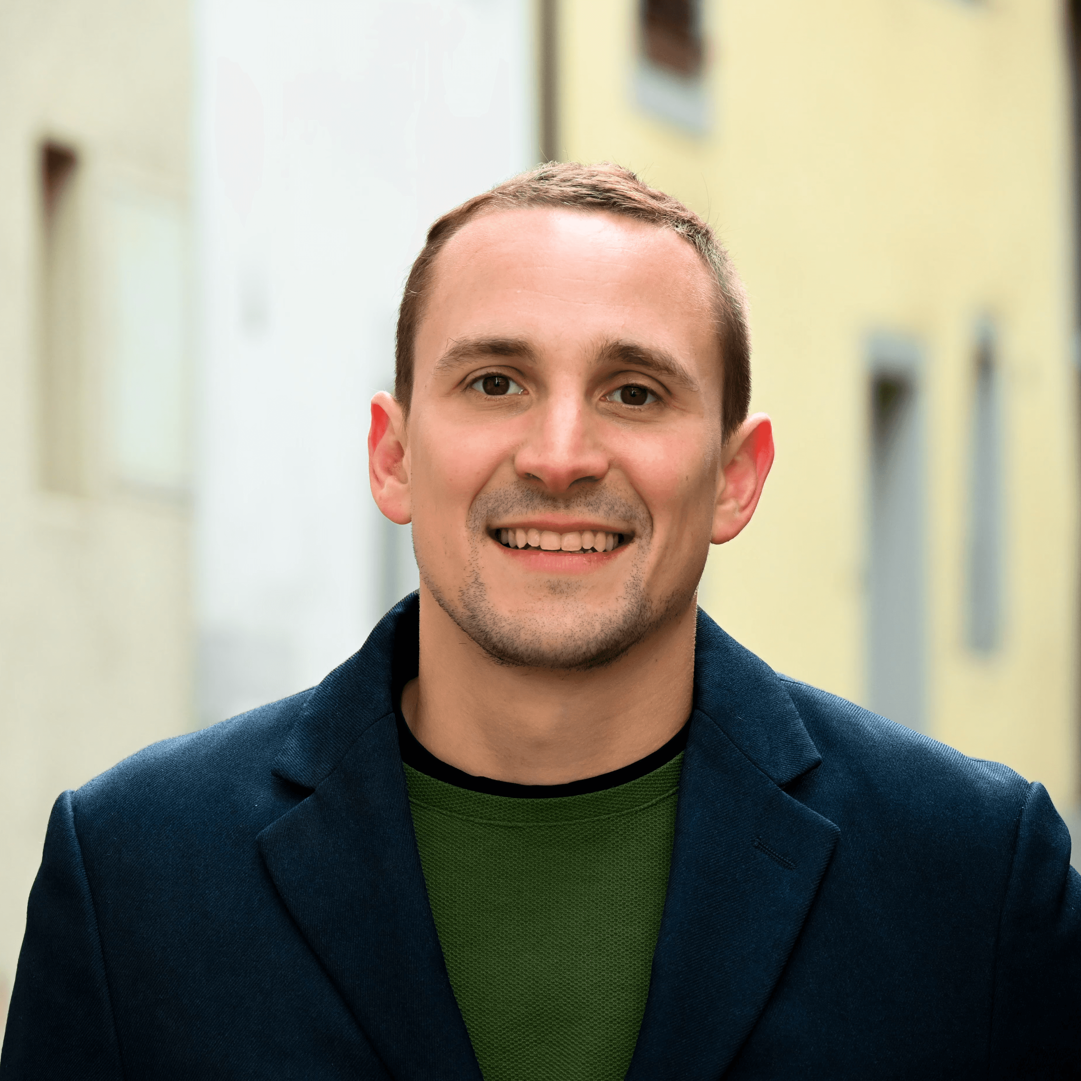 Joel de Buren in blazer smiling outdoors, with soft-focus urban background.