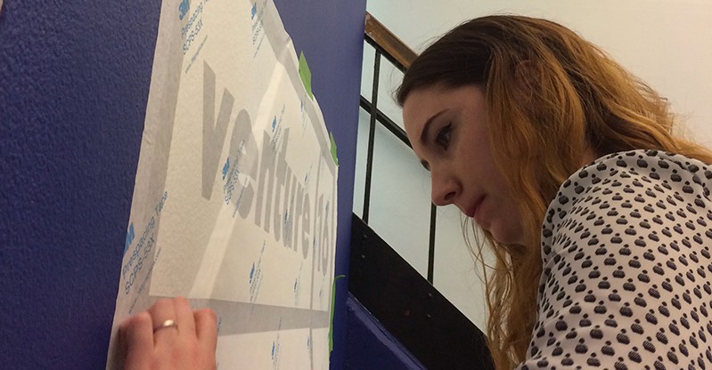 A woman sticking a venture 16 logo on a blue wall.