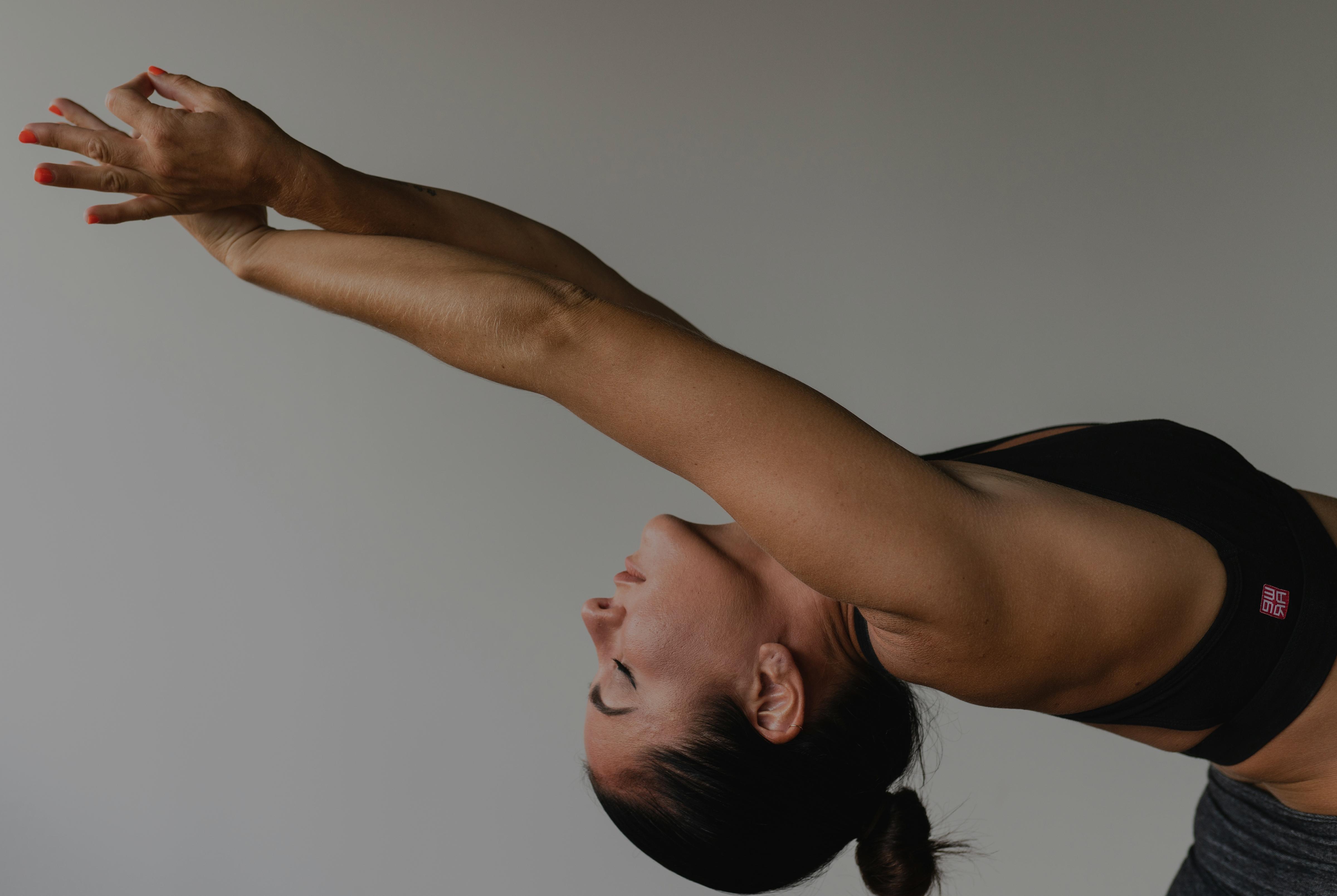 Woman performing a standing backbend stretch with arms extended overhead against a neutral studio background.