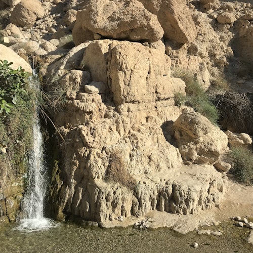 Small waterfall cascading over jagged rocks into a shallow pool, surrounded by dry, rocky terrain and sparse vegetation.