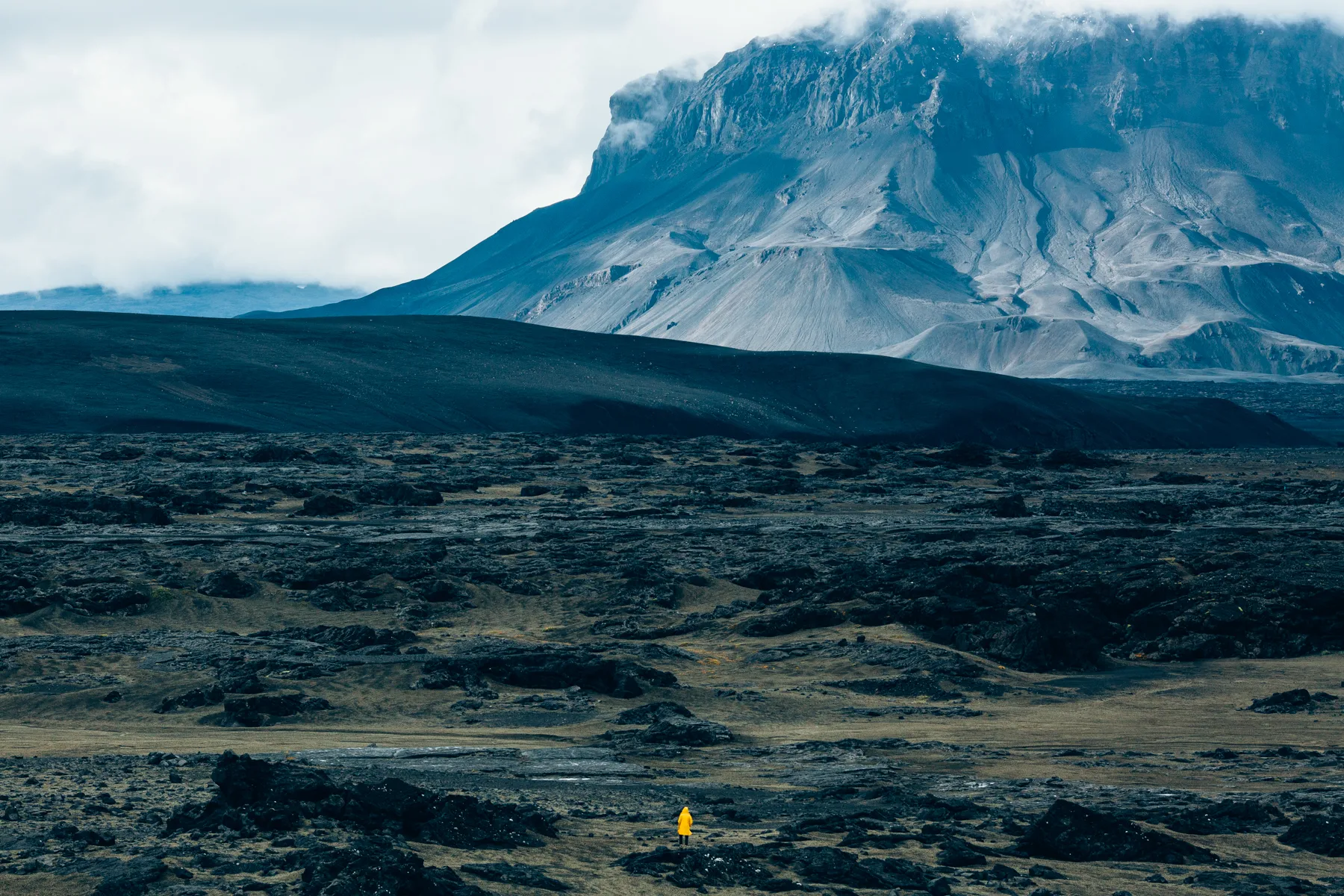 A snow-streaked mountain rising above a dark lava field, a tiny figure for scale