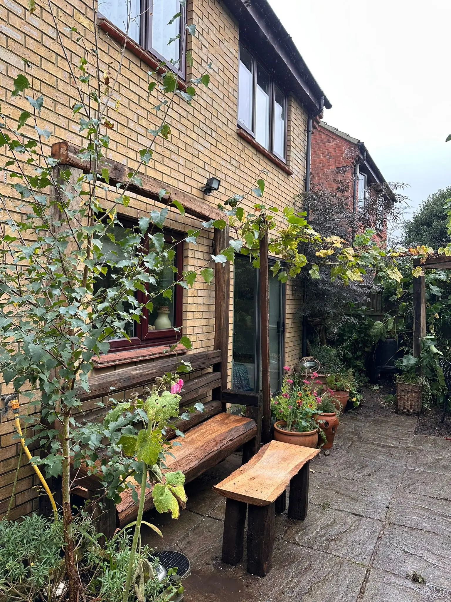 A cozy garden space featuring a wooden bench, plants, and a brick wall of a house under a cloudy sky.