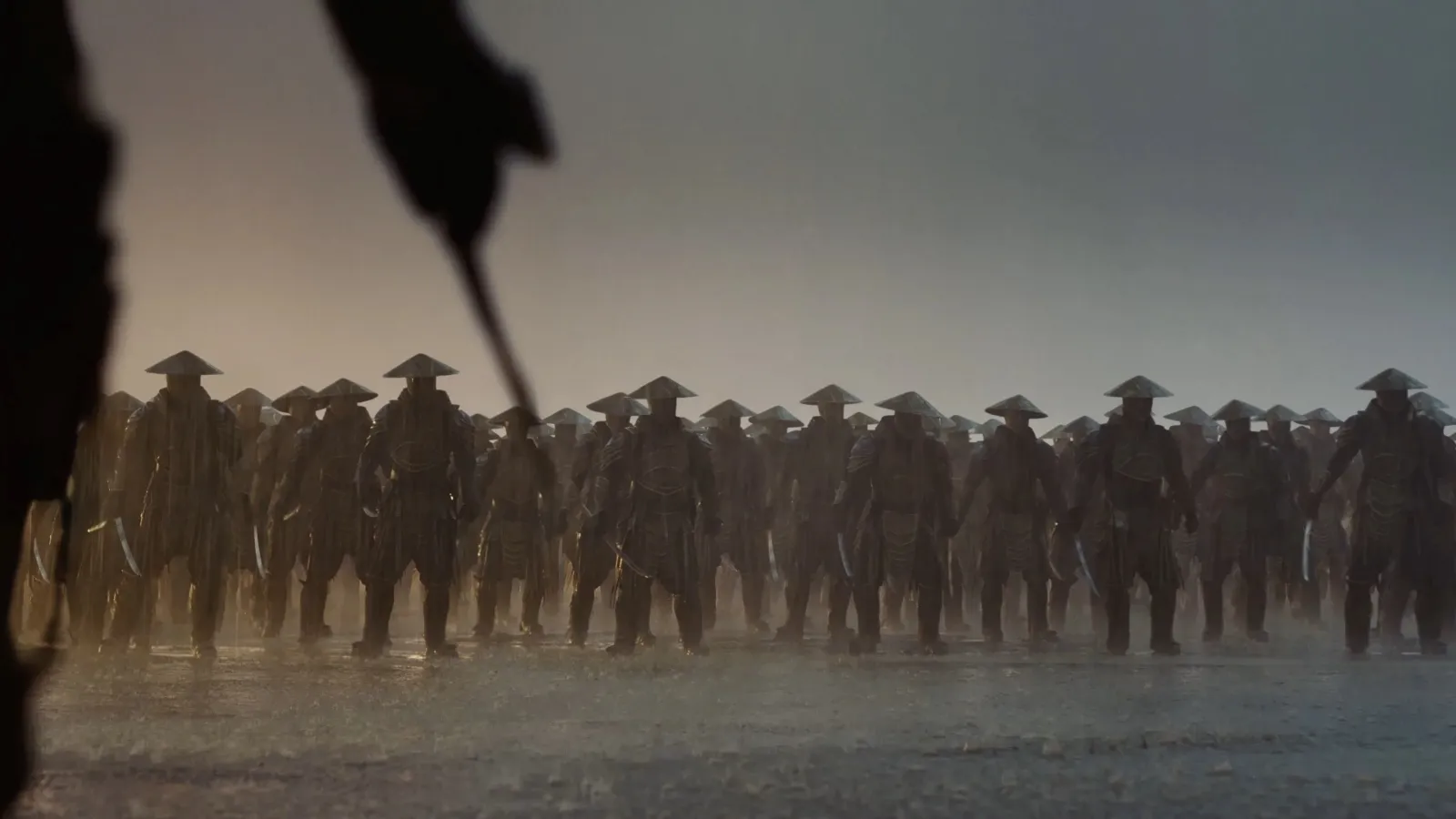 A large group of warriors with samurai style head wear, wearing traditional armour and conical helmets, stand in formation on a misty battlefield at dusk, ready for battle.