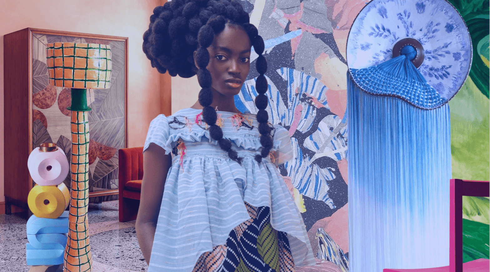 Imagem com estética afrofuturista e linguagem visual contemporânea. Ao centro, uma mulher negra com penteado marcante e vestida com roupas em tecidos mistos e volumosos. O ambiente é composto por objetos de design artístico e colorido — como esculturas geométricas, luminária com padrão quadriculado e painel de mosaico ao fundo. A cena transmite uma fusão entre cultura, arte e inovação.  O texto sobreposto diz: “Inovação e Cultura para além das Tendências” e “Beleza & estética 2026”, sinalizando o tema da imagem — uma palestra ou conteúdo sobre macrotendências de consumo, estética, comportamento e inovação cultural.  Essa imagem representa o cruzamento entre tendências de consumo, design afrofuturista, moda identitária e direções estéticas para o futuro, sendo ideal para conteúdos sobre beleza 2026, comportamento do consumidor, inovação cultural e tendências emergentes.