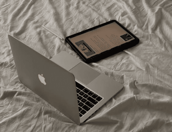 하얀 침구 위에 실버 노트북(맥북 맥락)과 태블릿 PC가 나란히 놓여 있는 모습입니다. 원룸 공간에서 인터넷을 이용해 콘텐츠를 즐기거나 작업하는 환경 상징합니다.