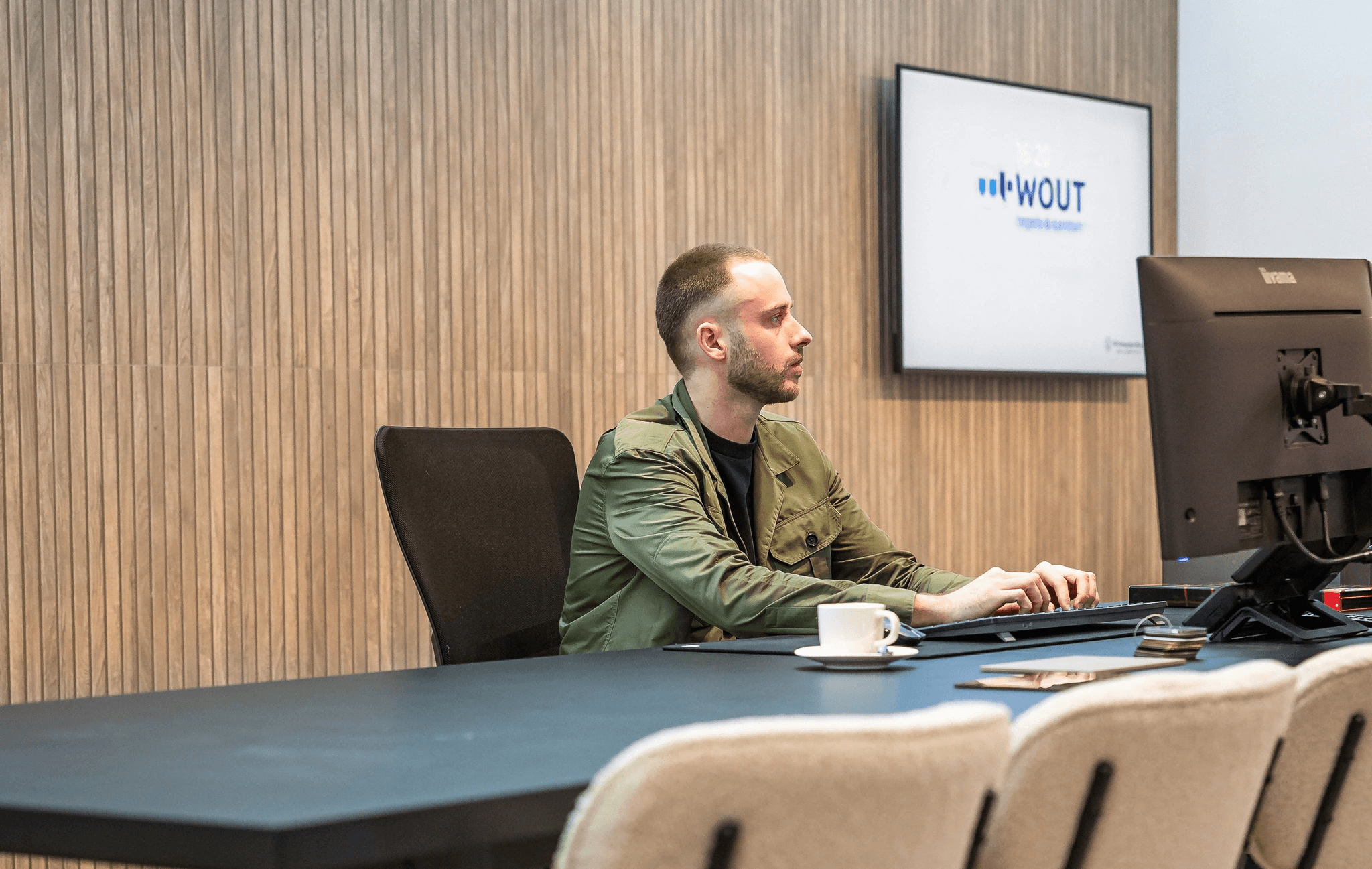 Young man in a green hoodie holding a yellow phone while seated at a table with a tablet.