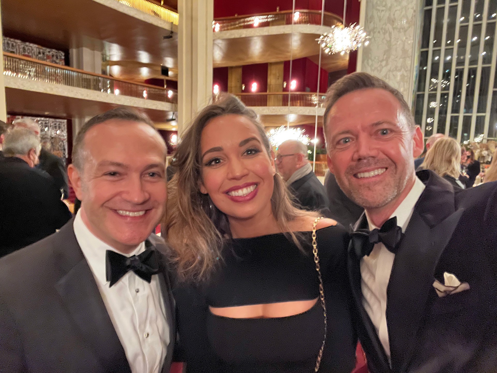 A beautiful woman in a black gown and dark hair stands flanked by two men in tuxedos, celebrating her success in an opera house