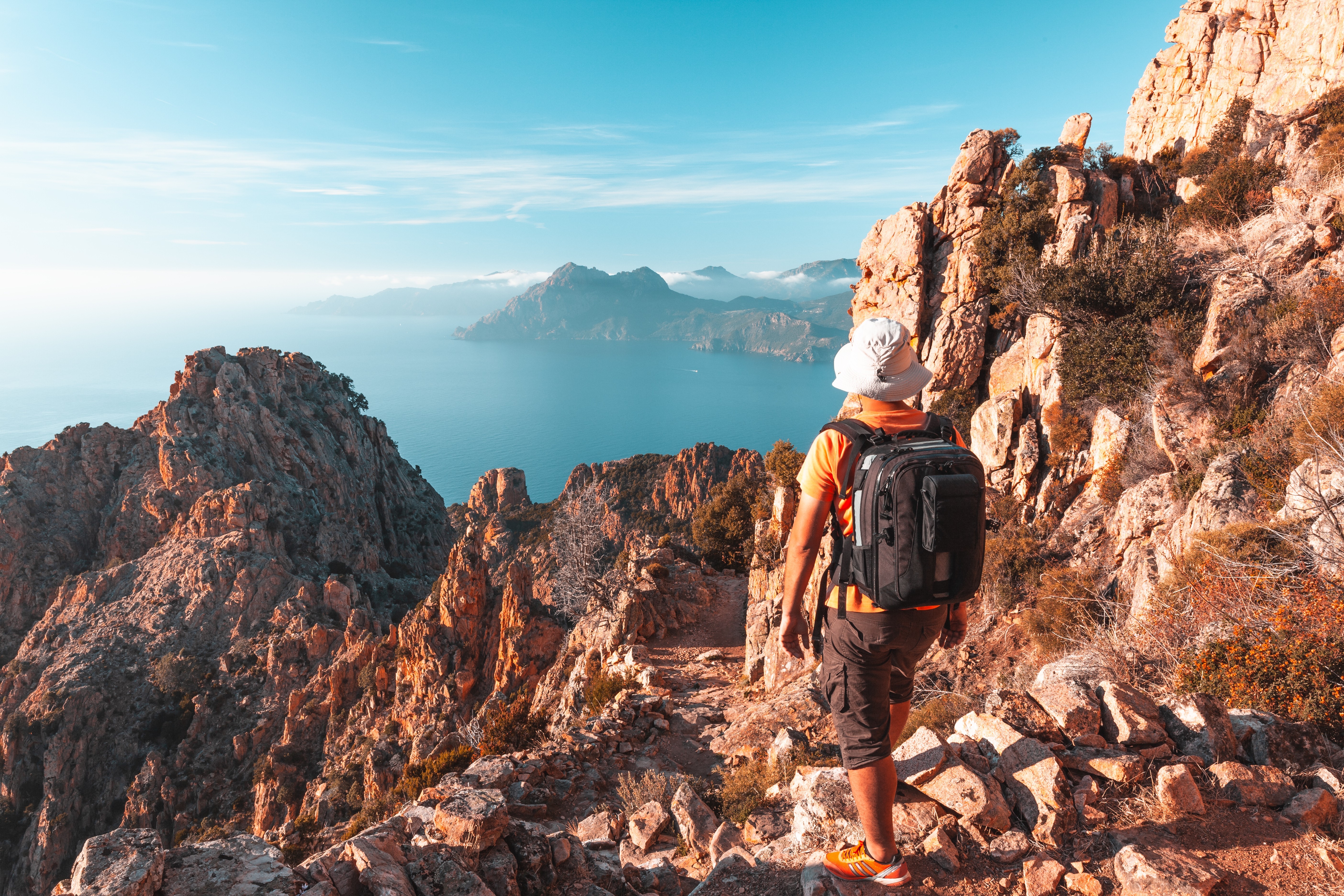 A hiker in shorts and a tshirt looks out across the turquoise Mediterannean Sea while hiking along a rocky path on Corsica's western coastline.