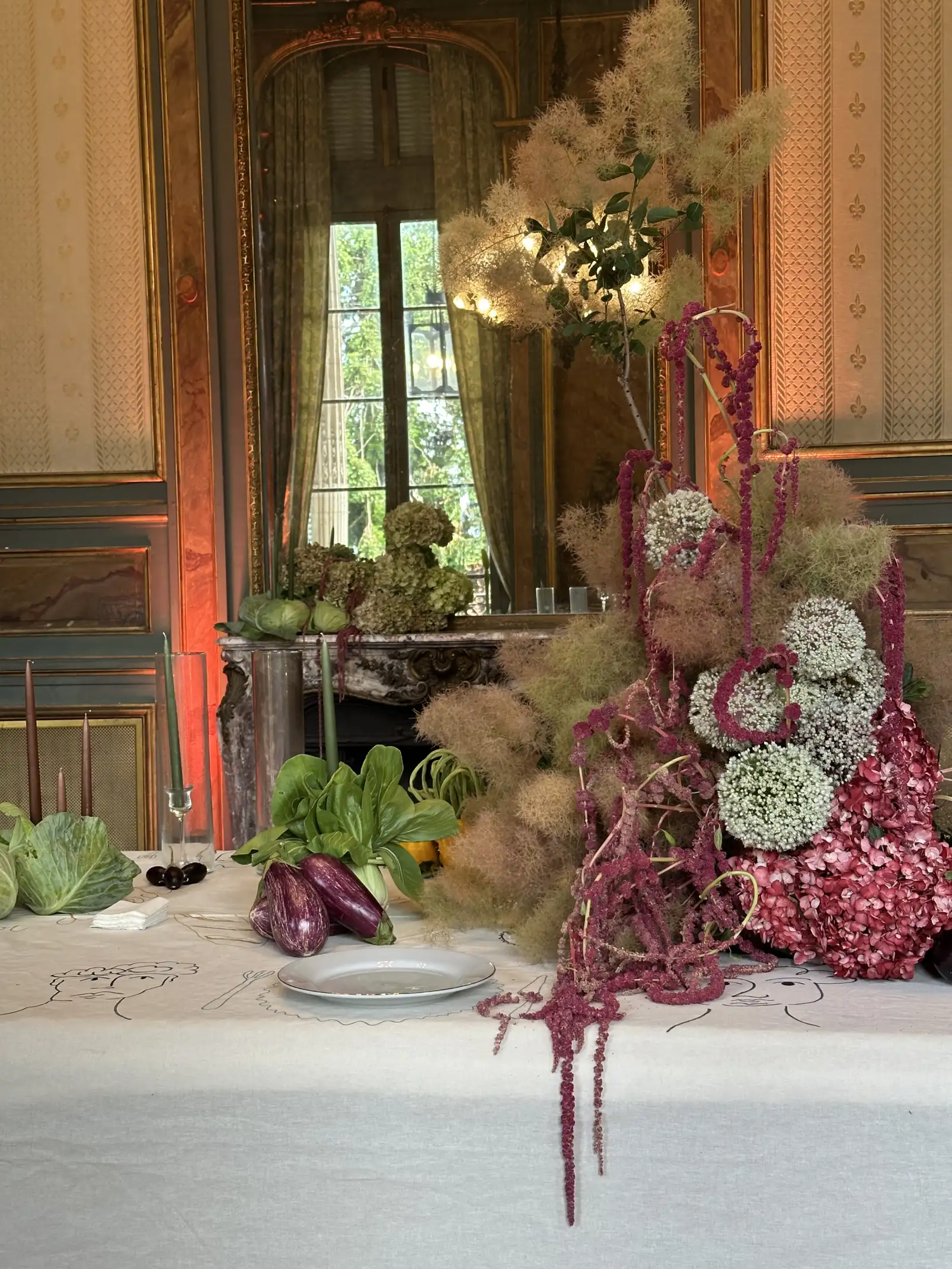 Mesa decorada con un centro de mesa floral de hortensias rosadas, flores blancas y amaranto rojo colgante, acompañado de berenjenas, repollo y hojas verdes sobre un mantel blanco bordado, en una sala elegante con espejo antiguo y detalles dorados