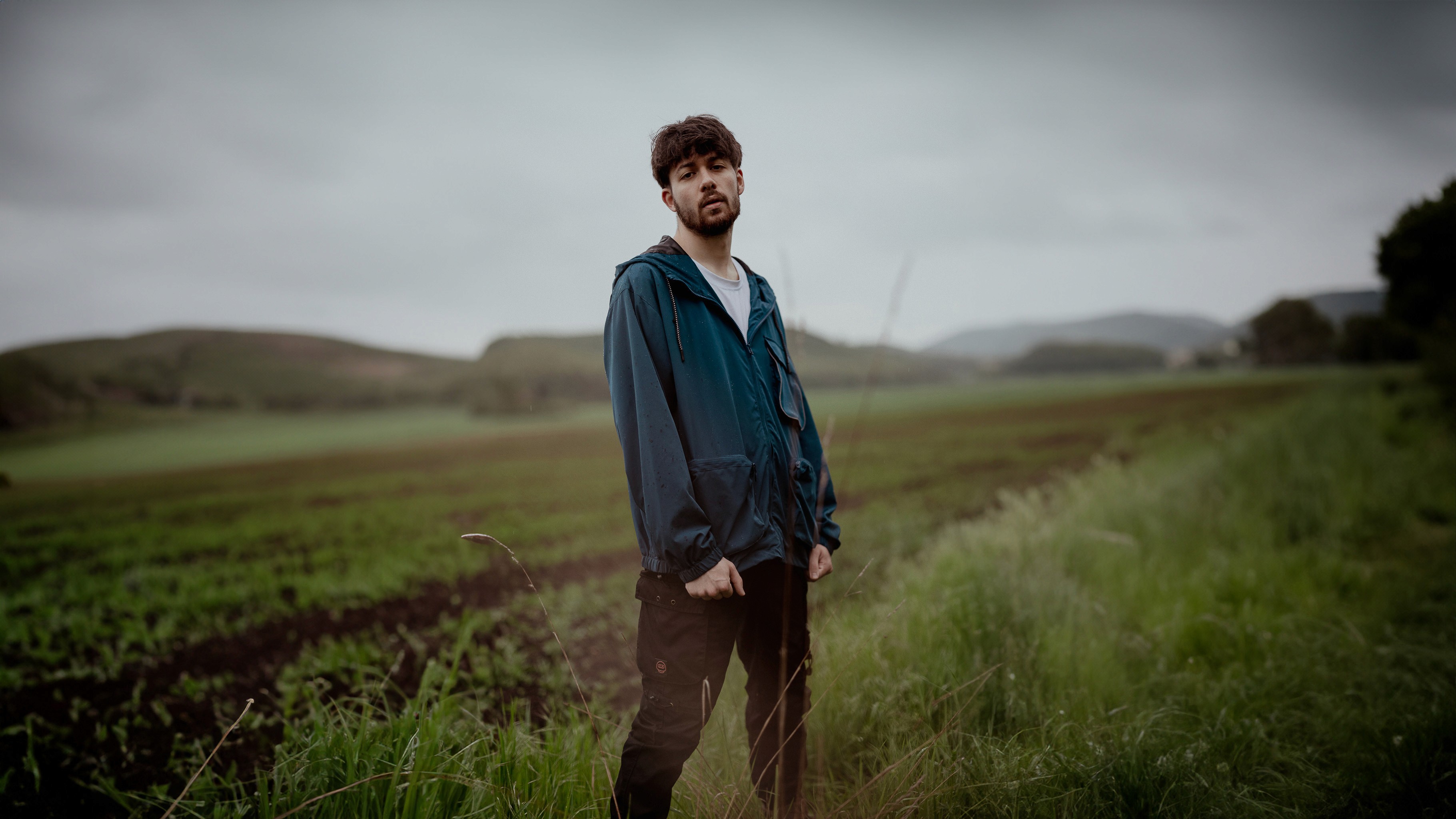 Egy fiatal férfi áll zöld dzsekiben, tavaszi mezőn és dombok között, borús ég alatt, természetközeli portré hangulattal.