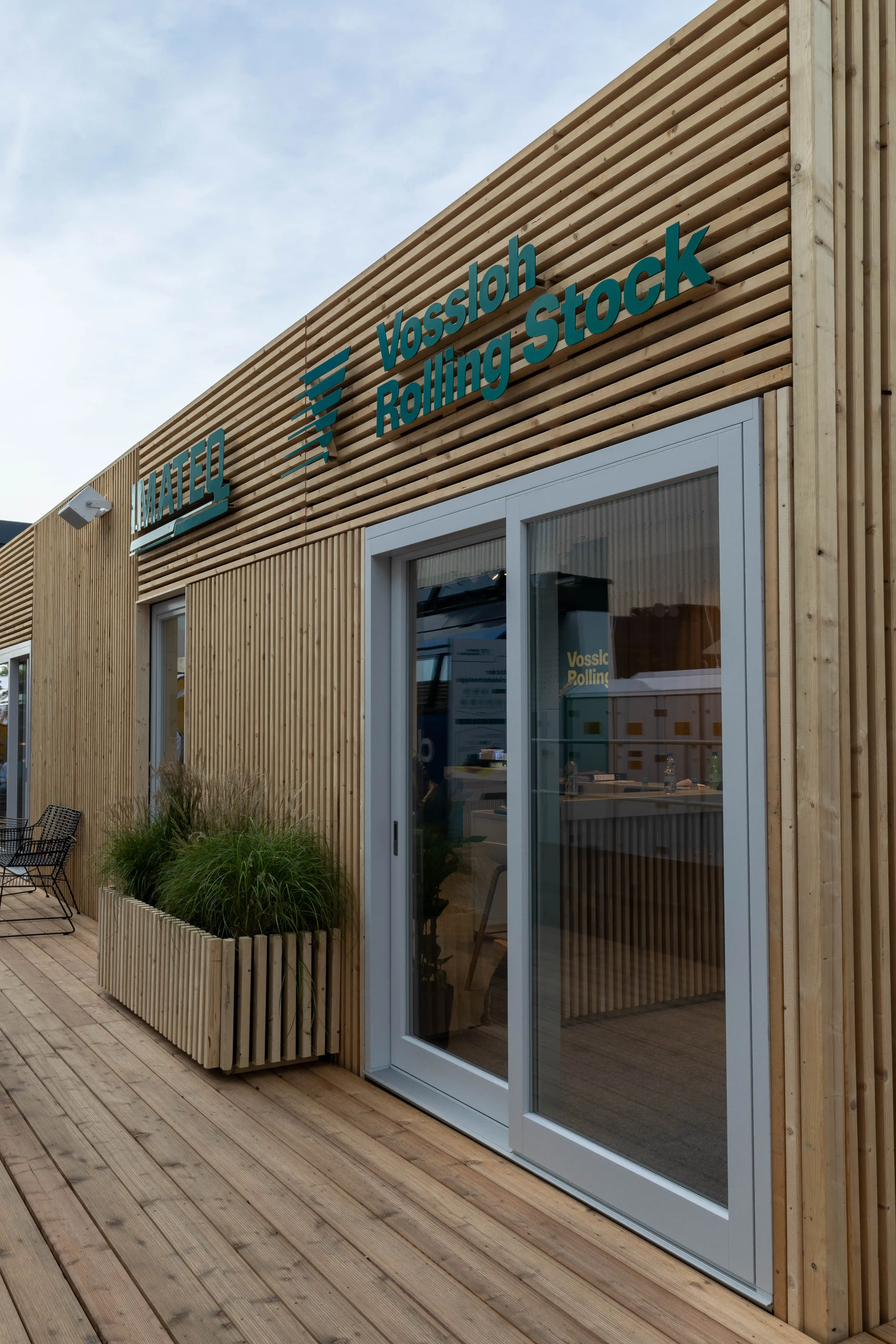 Entrance with sliding glass door in the larch-slat facade of the Vossloh Rolling Stock pavilion with wooden deck.