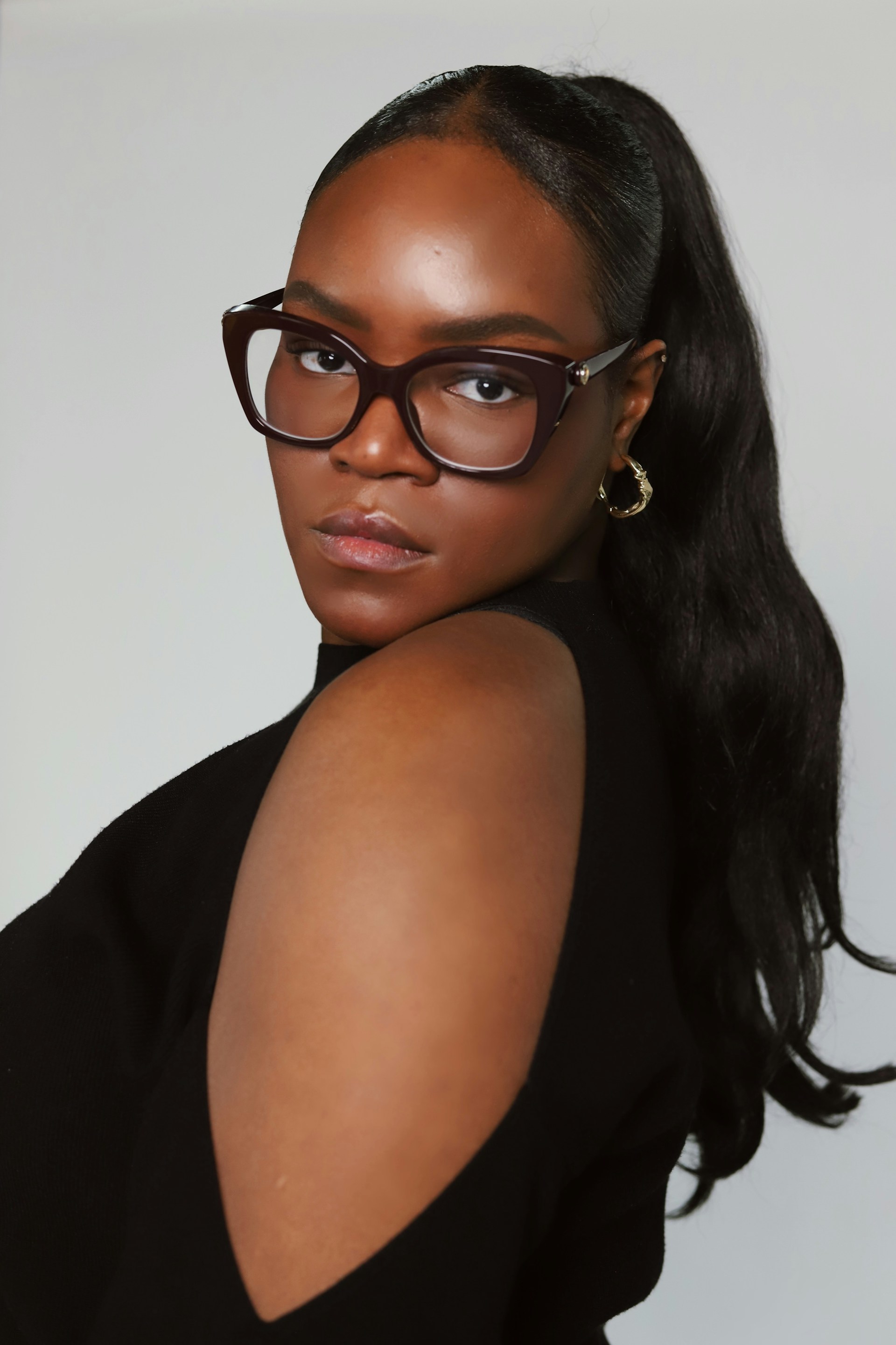 A close-up profile shot of the woman looking over her shoulder. She is wearing the same cat-eye glasses and gold hoop earrings, with her dark hair styled in a long, wavy ponytail.
