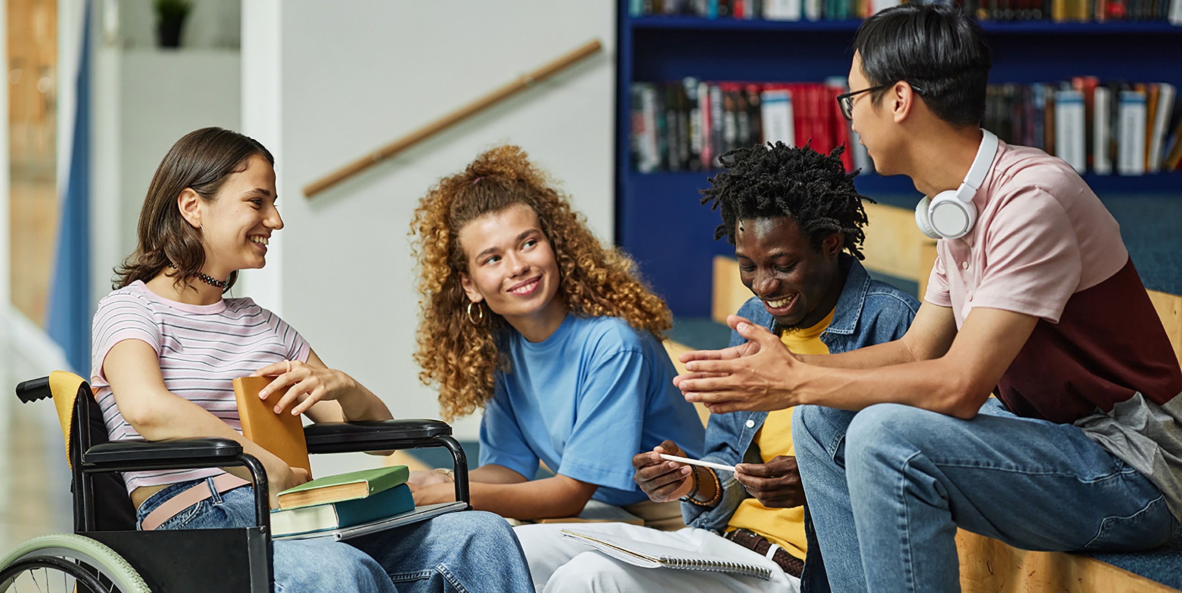 Four diverse students sit together in a bright library or study space, engaged in animated conversation and laughter. A young woman using a wheelchair sits on the left, alongside peers with books and study materials scattered around them. A bookshelf is visible in the background, depicting an inclusive educational environment where students of varying abilities collaborate and connect.