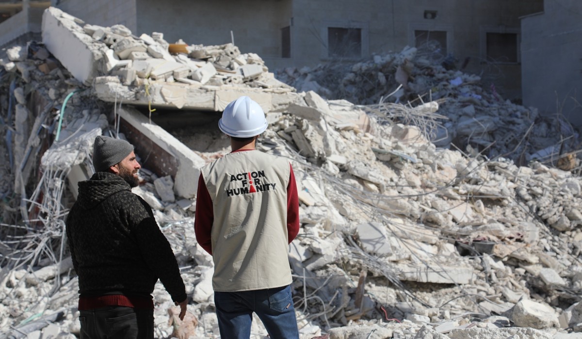 Two people watching over a demolished building. The person on the right wearing a vest with the logo of Action for Humanity on it