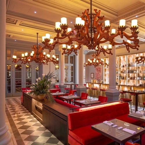 Isang eleganteng restawran na may mga pulang velvet na upuan, mararangyang chandelier, at sahig na may disenyong checkerboard. May bar sa likuran na may mga naka-display na bote.