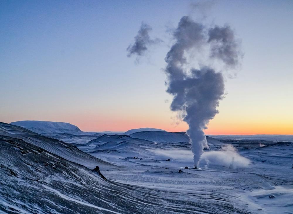 A winter landscape at sunrise with snow-covered hills and valleys. A large plume of steam rises from the ground, creating a serene, chilly atmosphere.