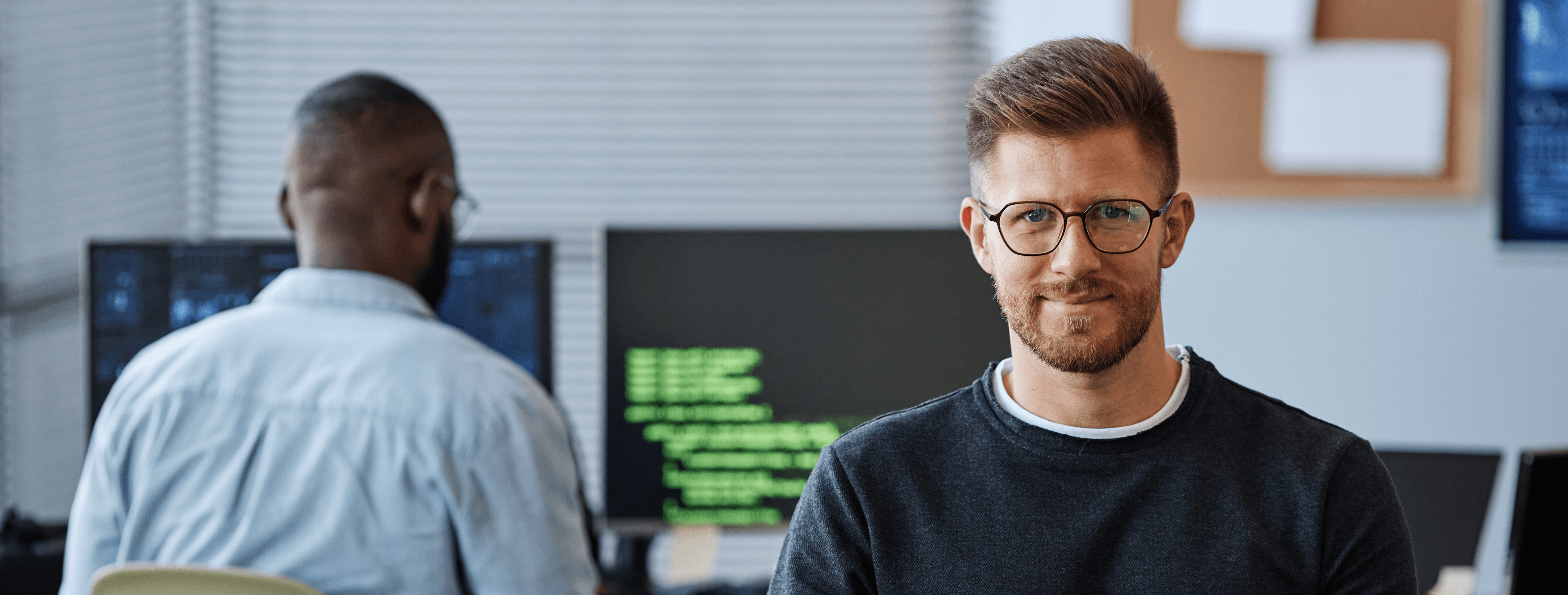Developer looking at the camera while a colleague writes code in the background, illustrating a professional engineering team.