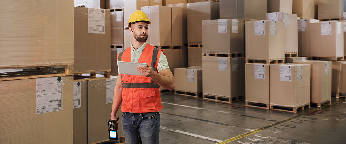 Worker in safety vest standing among stacked cardboard boxes in a warehouse.