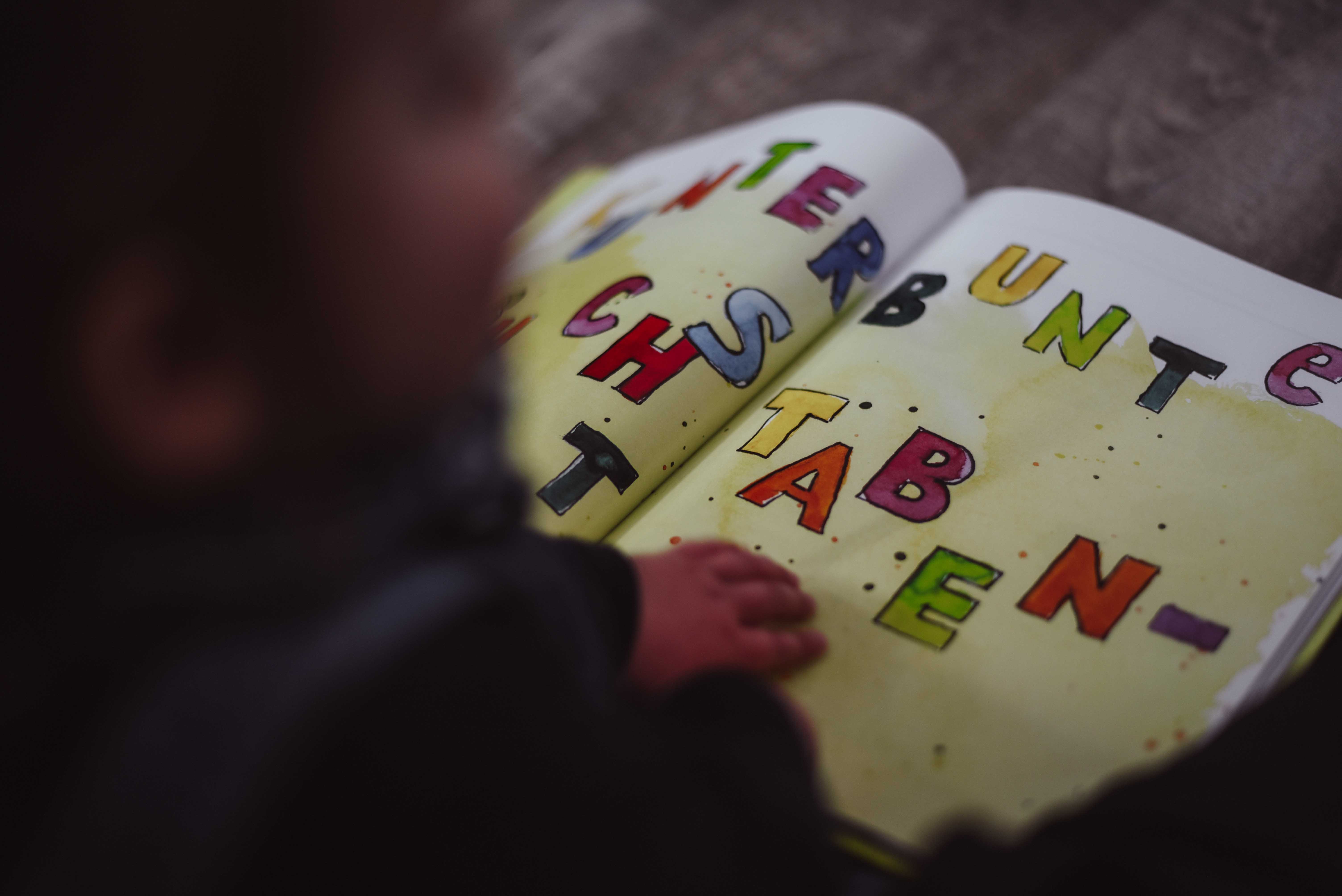 Detailaufnahme moderne Kitafotografie Gevelsberg: Ein Kind vertieft sich in ein farbenfrohes Buch mit der Aufschrift „Bunte Buchstaben“, fotografiert von PK photography 4 Kids NRW.