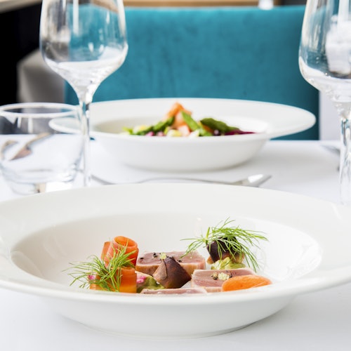 Elegantly plated dish with sliced meat, carrots, and greens on a white table, set with wine glasses and another dish in the background.