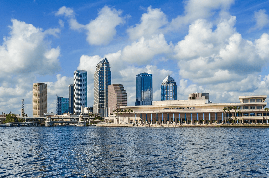 A waterfront city skyline with modern high-rise buildings under a bright blue sky filled with scattered white clouds. In the foreground is a body of water, and along the shoreline stands a large convention center building with columns and palm trees lining the walkway. The scene conveys a sunny, coastal urban setting.