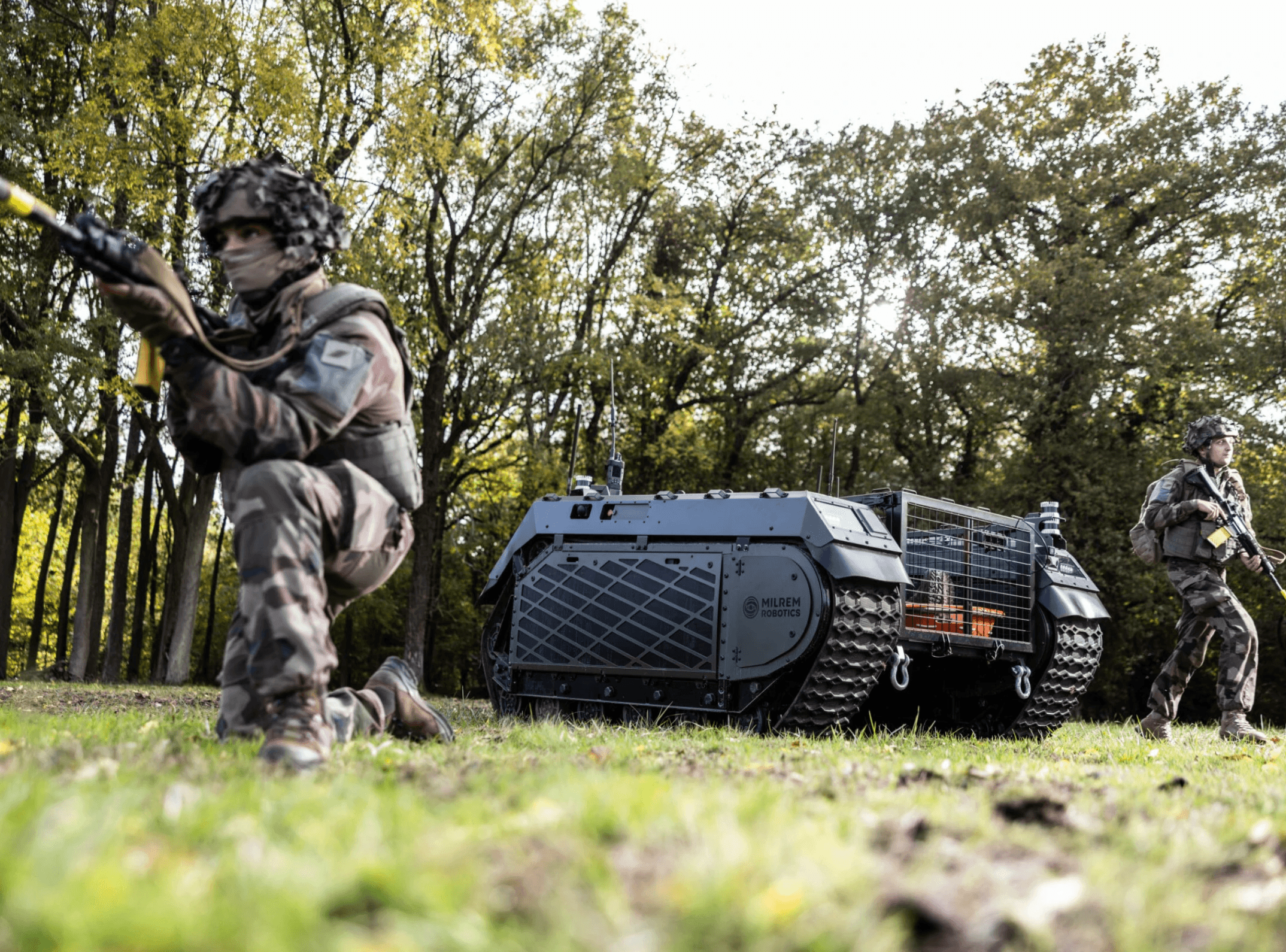 In a forested setting, two soldiers in full camouflaged gear are positioned in front of an unmanned, tracked military vehicle as they hold rifles and survey their surroundings, highlighting a scenario of modern, technology-supported defense operations.